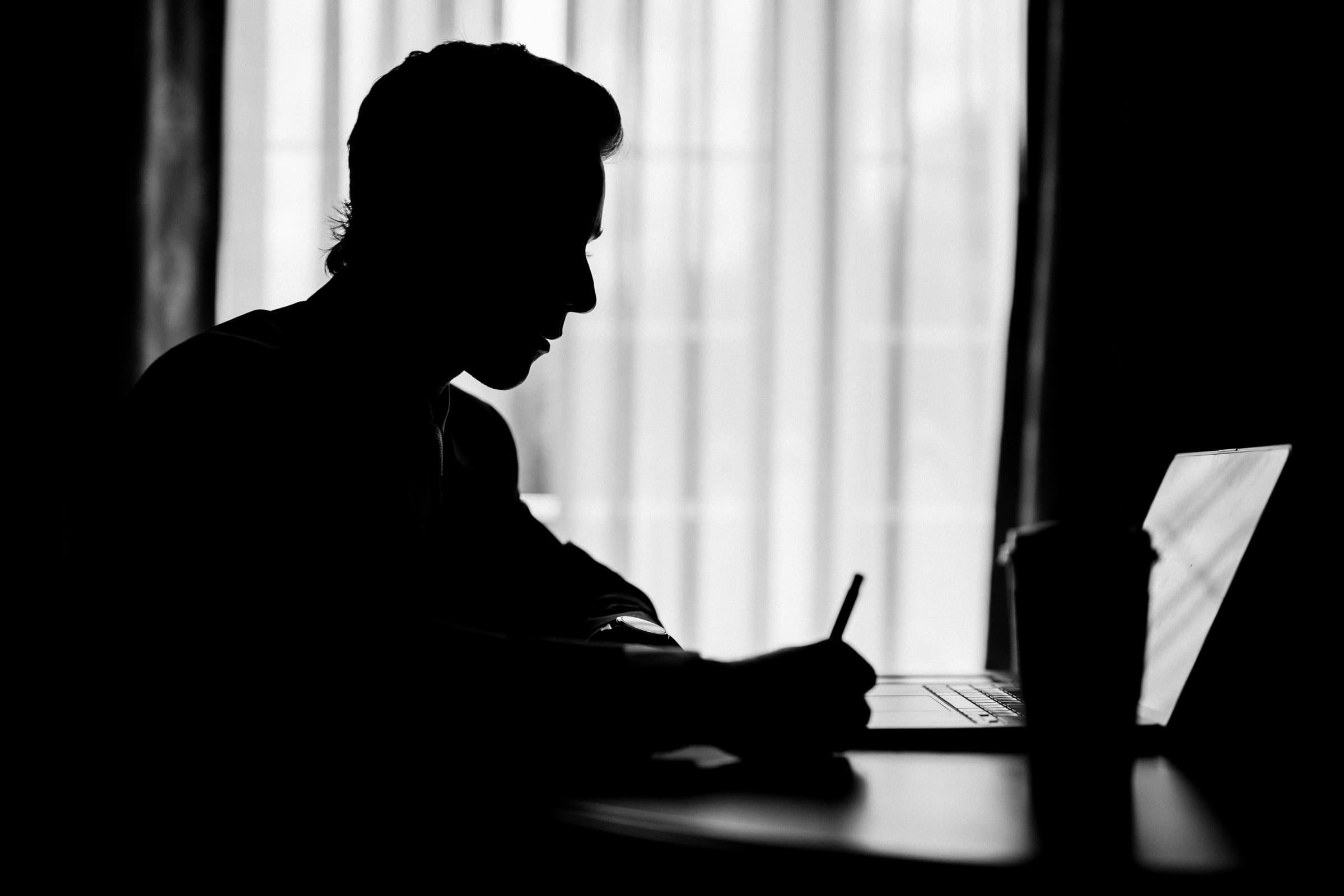 duke university wedding photography - sarah & case Silhouette of a person working at a desk with a laptop, backlit by window curtains. The person holds a pen, and a cup is visible on the desk, crafting an aura of peaceful focus reminiscent of Duke University’s library—ideal for wedding photography that captures intimate study moments.