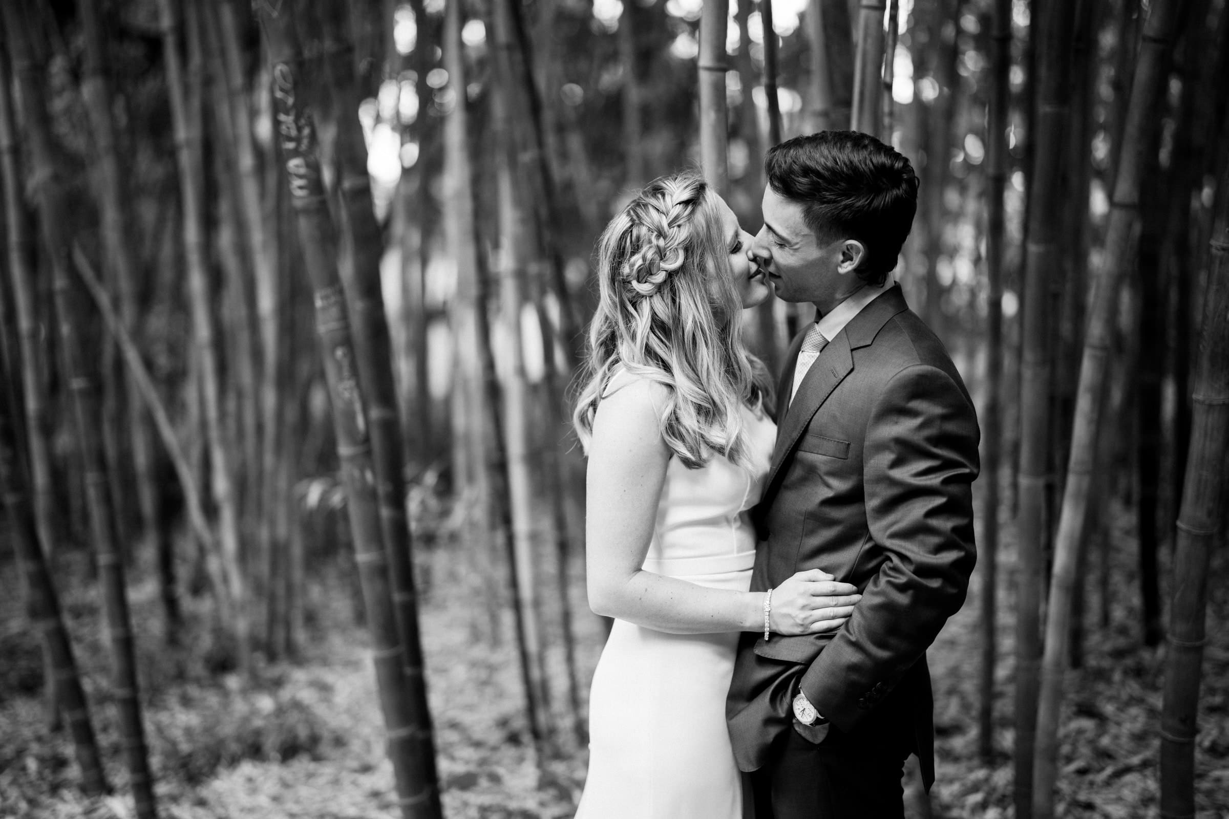 A black and white photo by a Raleigh wedding photographer captures a couple in formal attire amidst a bamboo grove. The woman, in a dress with braided hair, and the man, in a suit, gaze at each other with smiles, creating an enchanting romantic atmosphere.