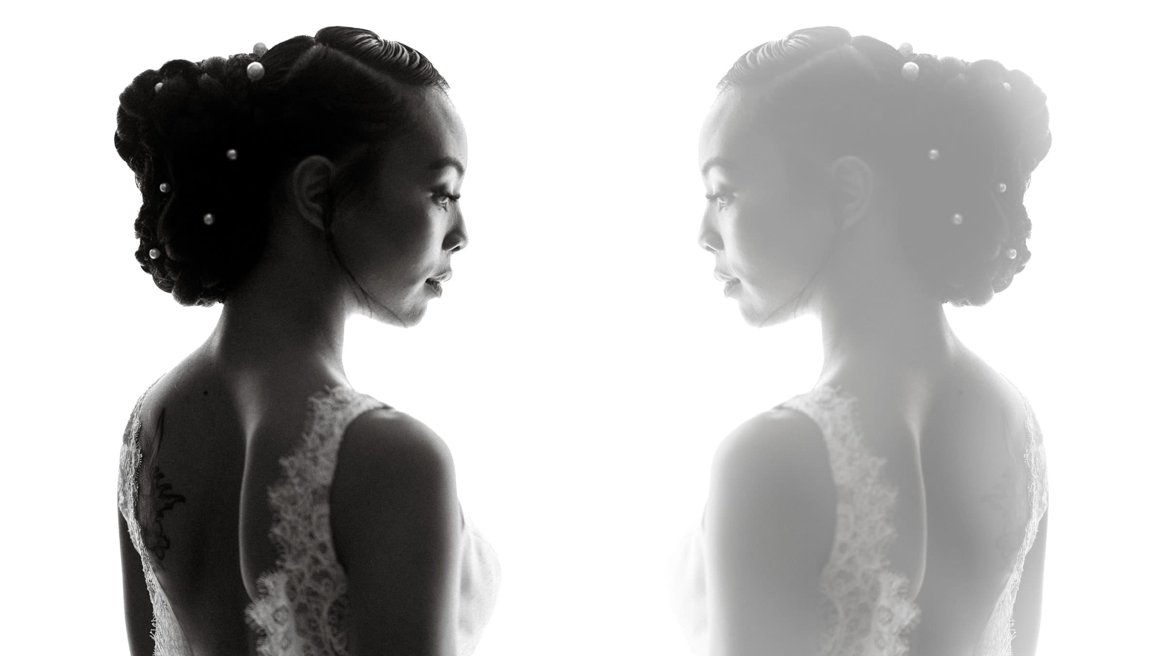 umstead wedding photography - kayla & deandre A black and white image captures a bride from an Umstead wedding with an elegant updo adorned with pearls. She gazes into a mirror, creating a reflective silhouette. The lace details on her dress enhance the serene and timeless atmosphere.