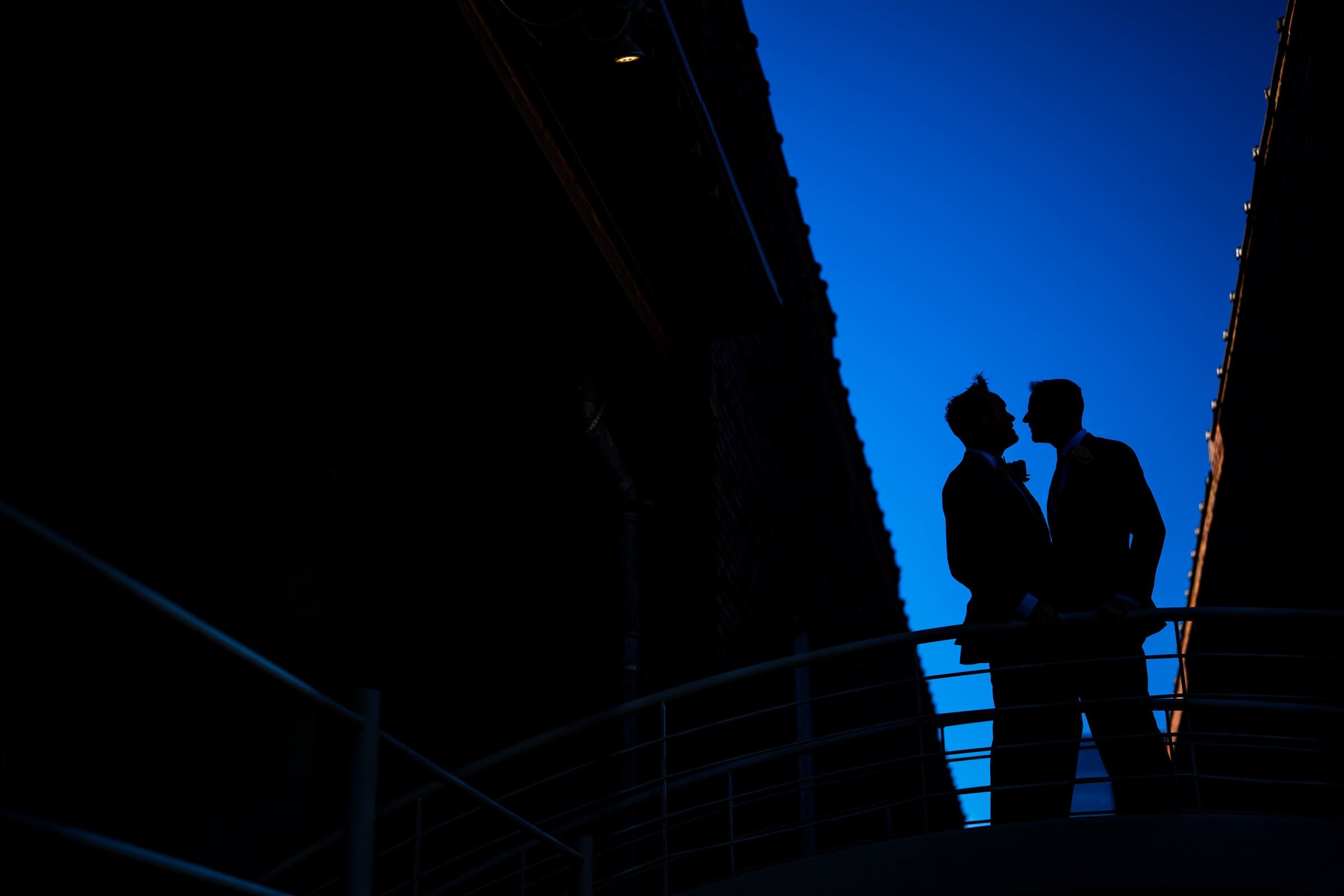 Silhouetted against a deep blue sky, two people in formal attire share a moment on a balcony at Union Station Raleigh, their figures outlined by the contrasting light. A railing curves in front of them, while dark structures frame this perfect wedding photography scene on both sides.