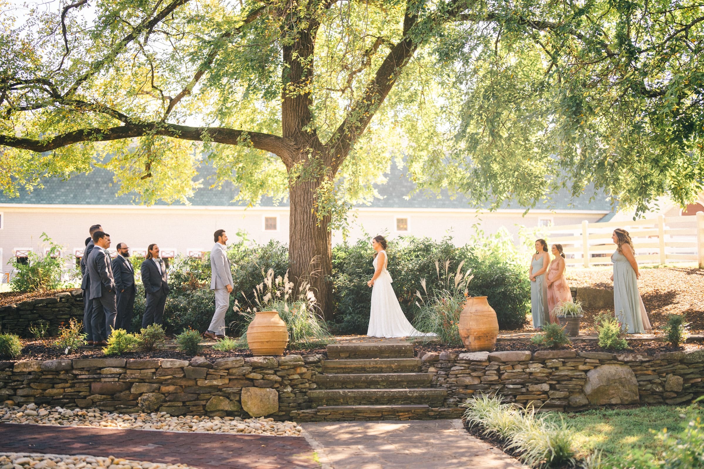 historic wakefield barn wedding - logan & rhett At a Wakefield Barn wedding, the ceremony unfolds outdoors under a grand tree. The bride and groom stand on stone steps, flanked by groomsmen to the left and bridesmaids to the right, all enveloped by lush greenery and elegant urns.