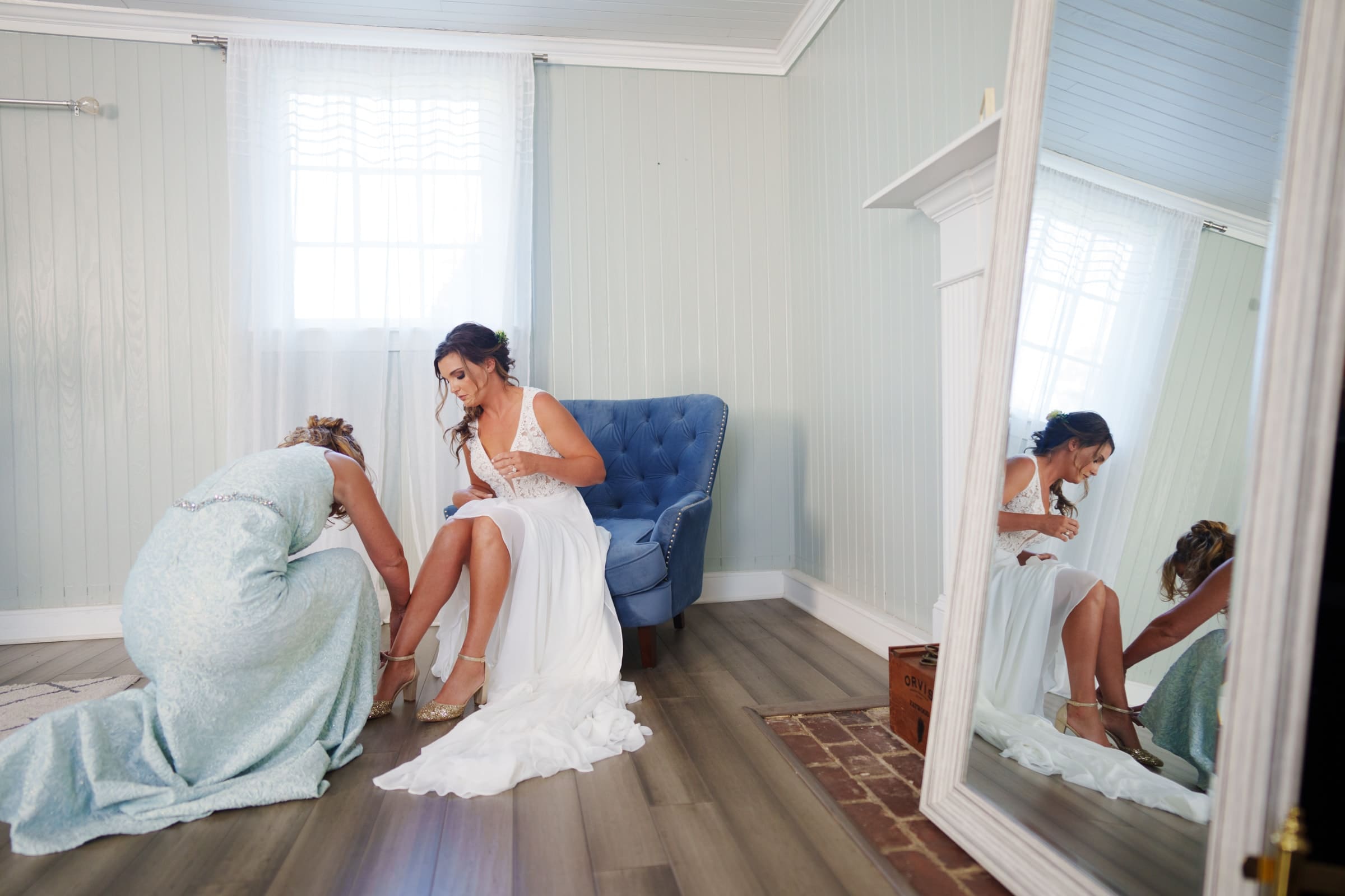 historic wakefield barn wedding - logan & rhett In a light-filled room at Wakefield Barn, a bride in a white dress sits on a blue chair as a woman assists with her shoe. Wooden floors gleam under the softly lit window, and a large mirror reflects the serene space with its light-colored walls.