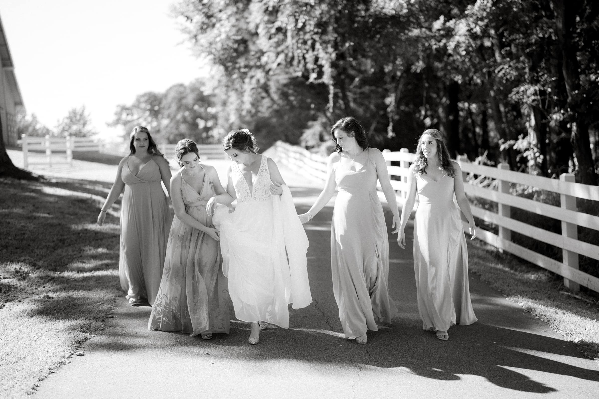 historic wakefield barn wedding - logan & rhett A black and white photo captures the charm of a Wakefield Barn wedding, with five women in long dresses strolling down a tree-lined path. The central woman, in a white dress, playfully lifts its hem while the others walk alongside her past the rustic white fences.