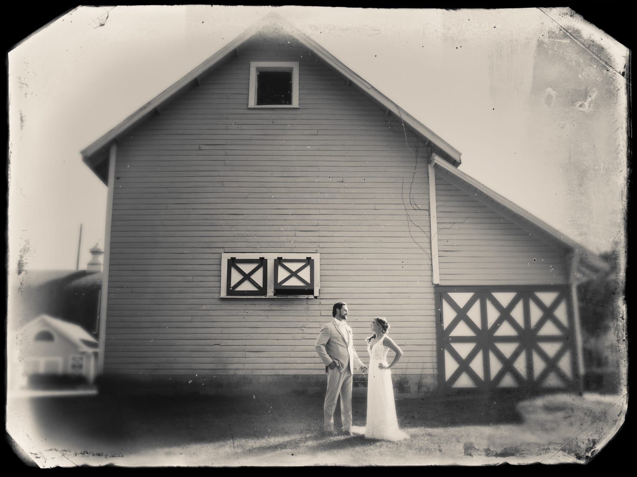 historic wakefield barn wedding - logan & rhett A sepia-toned photo captures a bride and groom facing each other at their Wakefield Barn wedding, framed by the rustic charm of crossed wooden doors and weathered wood siding. The groom's light suit contrasts beautifully with the bride's elegant white dress.