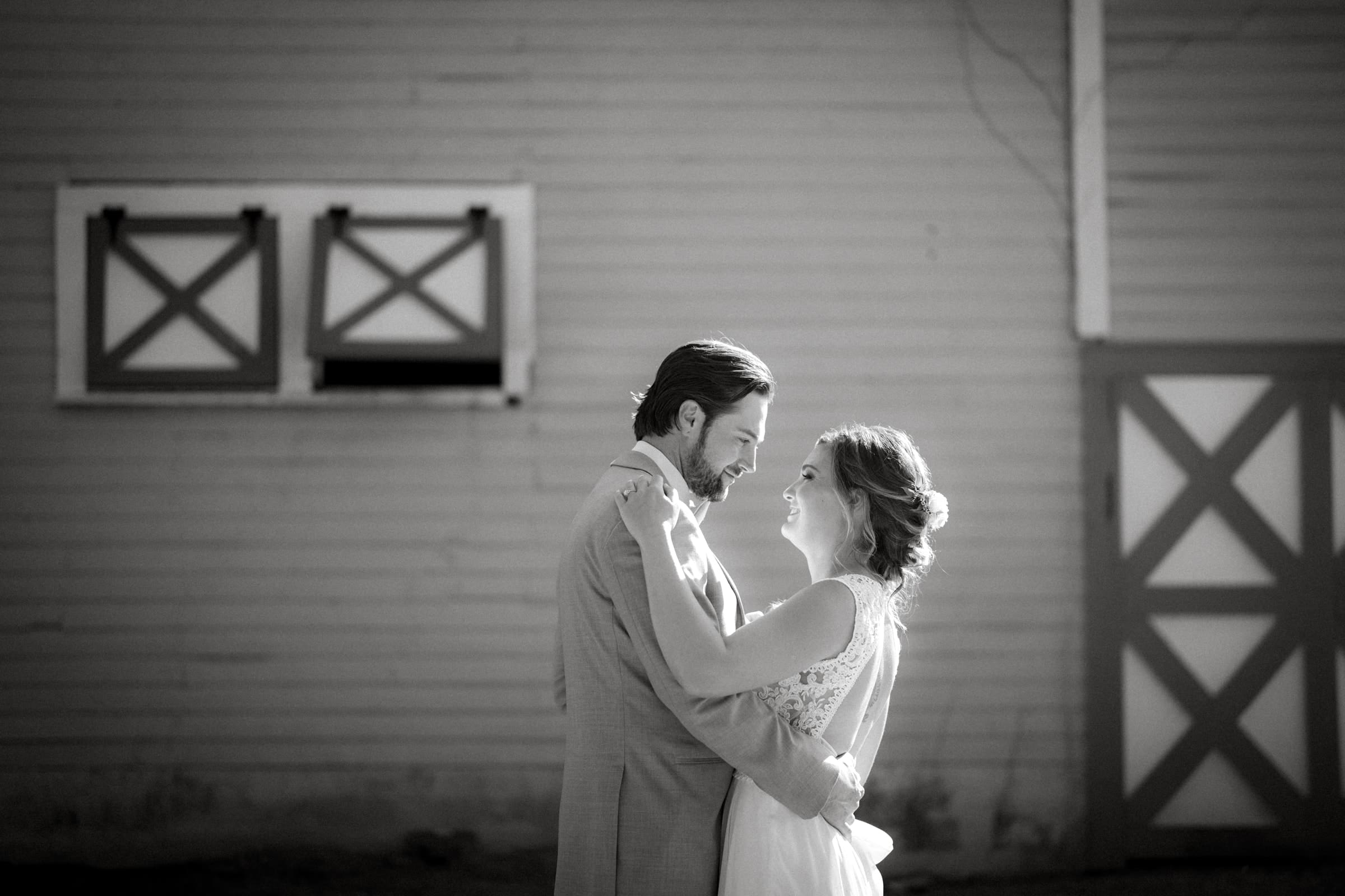 historic wakefield barn wedding - logan & rhett In this timeless black and white photo, a couple stands lovingly in front of the iconic Wakefield Barn. The groom, dapper in his suit, and the bride, radiant in her wedding dress, gaze into each other's eyes with gentle smiles, capturing the essence of their special day.