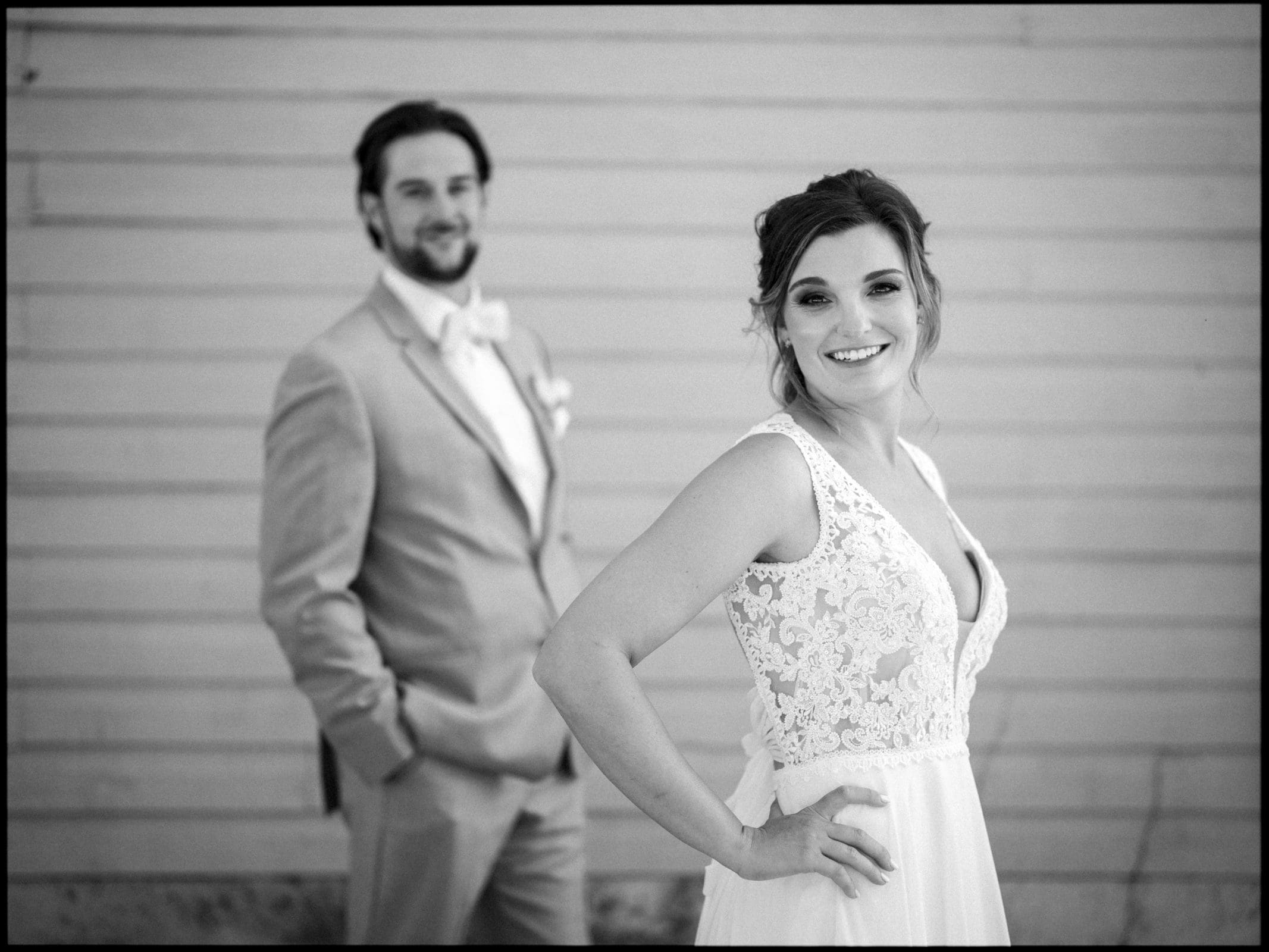 historic wakefield barn wedding - logan & rhett A bride and groom pose for a Wakefield Barn wedding photo. The bride is in the foreground, smiling, wearing a lace-trimmed gown. The groom, slightly blurred, is in a suit with a bow tie, standing behind her against a light-colored wall.