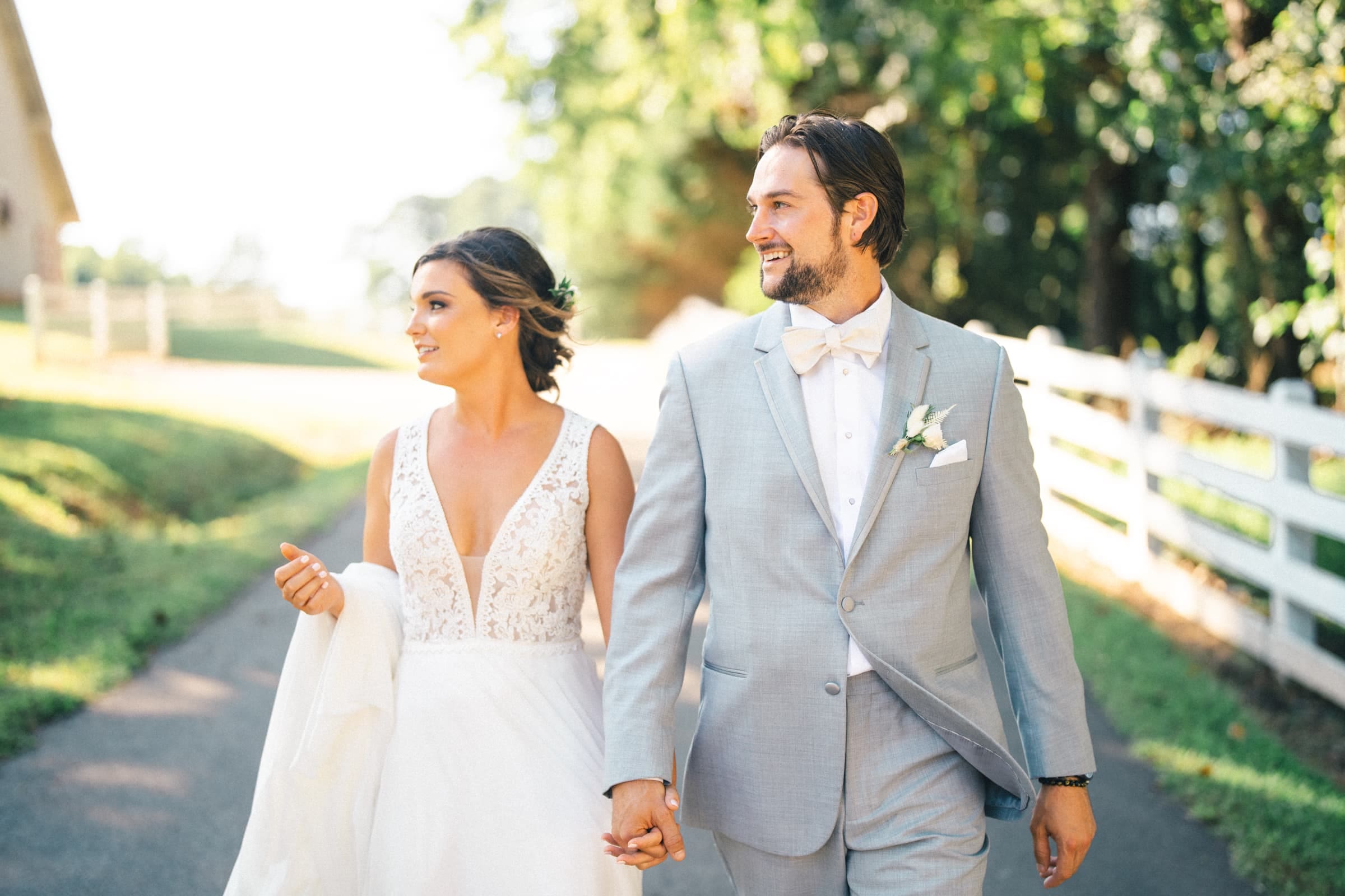 historic wakefield barn wedding - logan & rhett A bride and groom walk hand in hand along a sunlit path at Wakefield Barn, surrounded by greenery and a white fence. The bride wears a white gown, while the groom sports a light gray suit with a white bow tie and boutonniere.