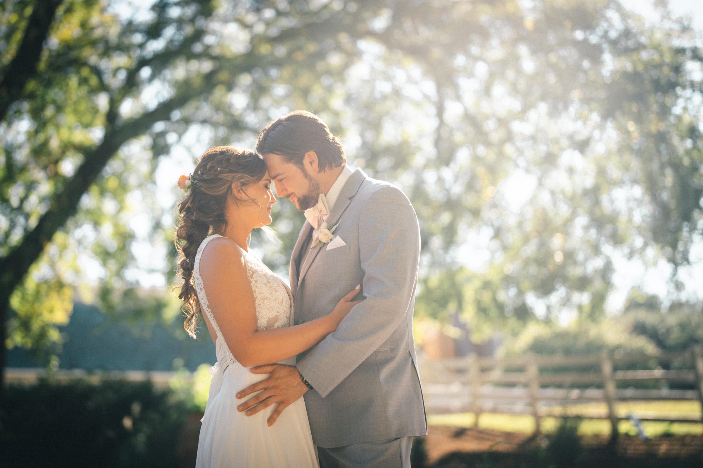 A bride and groom stand closely together, smiling under the warm sunlight at their Wakefield Barn wedding. They are outdoors, surrounded by trees and a wooden fence. The bride wears a white dress, and the groom is in a gray suit. It is a peaceful, romantic scene.