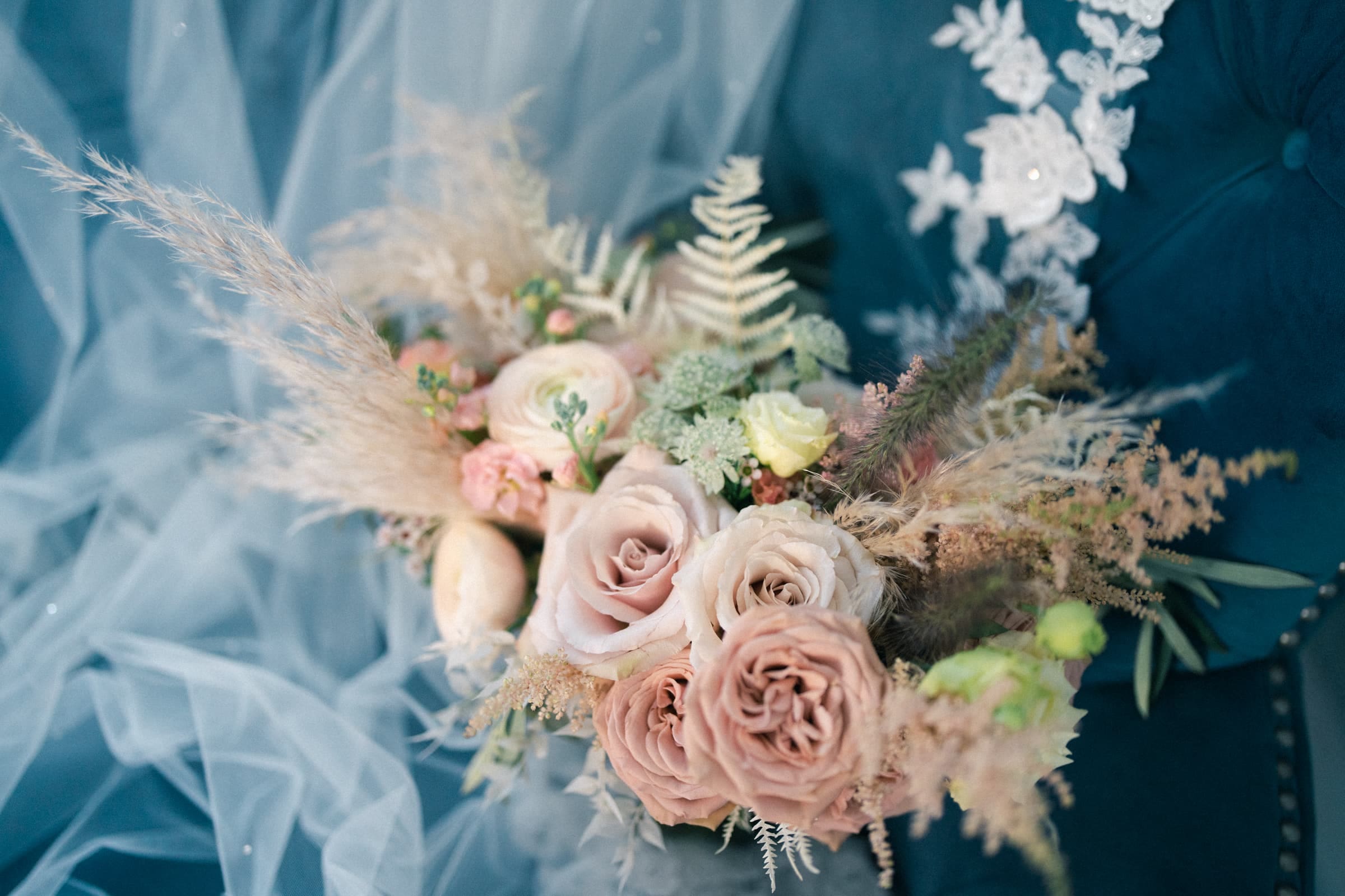 historic wakefield barn wedding - logan & rhett A bridal bouquet featuring blush and cream roses, surrounded by greenery and pampas grass, rests on a blue fabric with white lace detail, perfectly capturing the enchanting charm of a Wakefield Barn wedding.