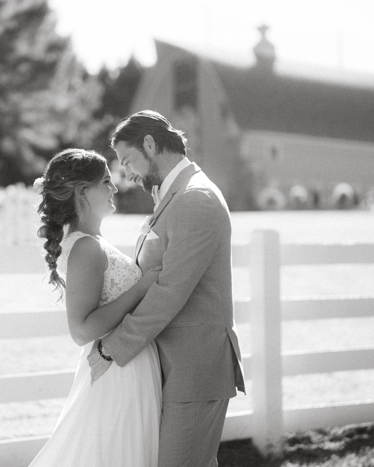 historic wakefield barn wedding - logan & rhett A bride and groom stand smiling at each other in front of a white fence at Wakefield Barn. The bride wears a long white dress while the groom sports a light-colored suit. The setting is bright and outdoors, perfectly capturing the romantic essence of a Wakefield Barn wedding.
