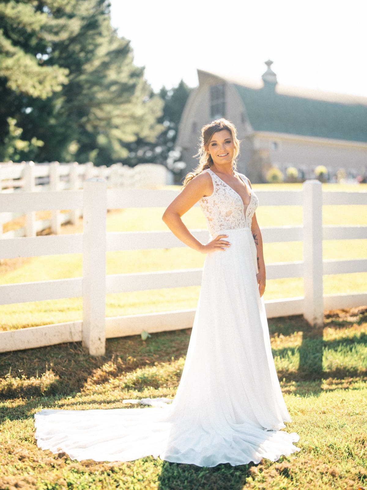 historic wakefield barn wedding - logan & rhett A bride in a white gown stands gracefully before a white fence, with the picturesque Wakefield Barn and lush trees silhouetted in the background. The sunny day casts enchanting shadows on the vibrant green grass.