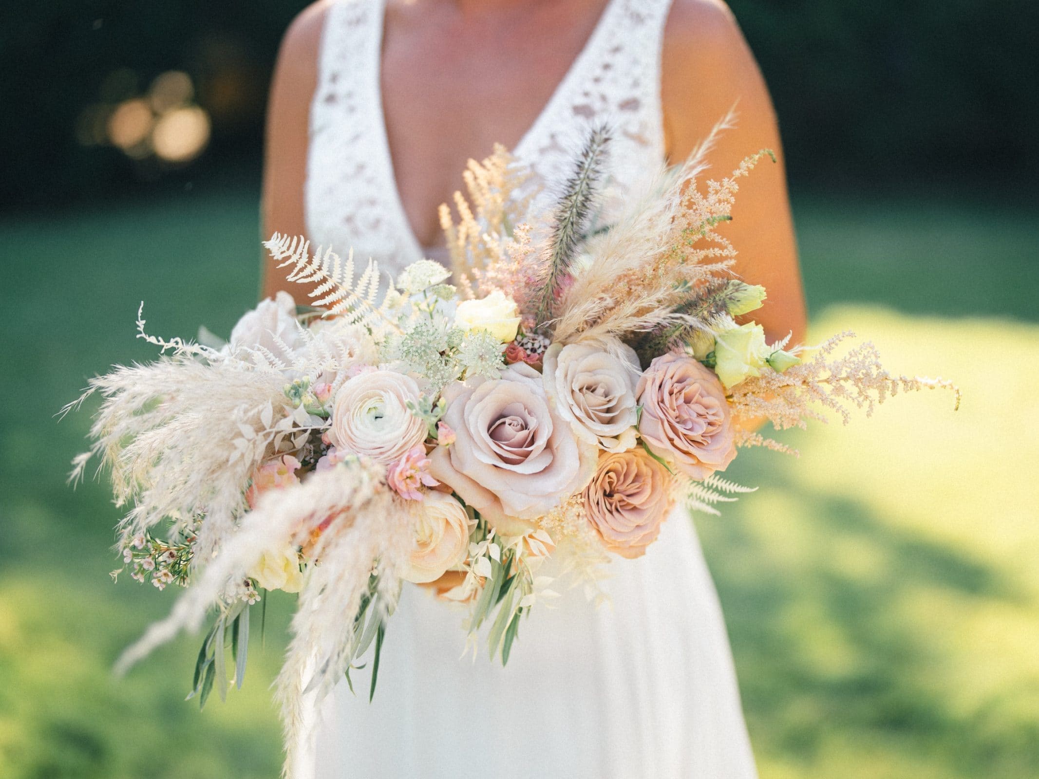 historic wakefield barn wedding - logan & rhett A person in a white dress holds a bouquet featuring pastel roses, peonies, pampas grass, and delicate ferns against the blurred greenery of a Wakefield Barn wedding. The bouquet exudes elegance and softness.