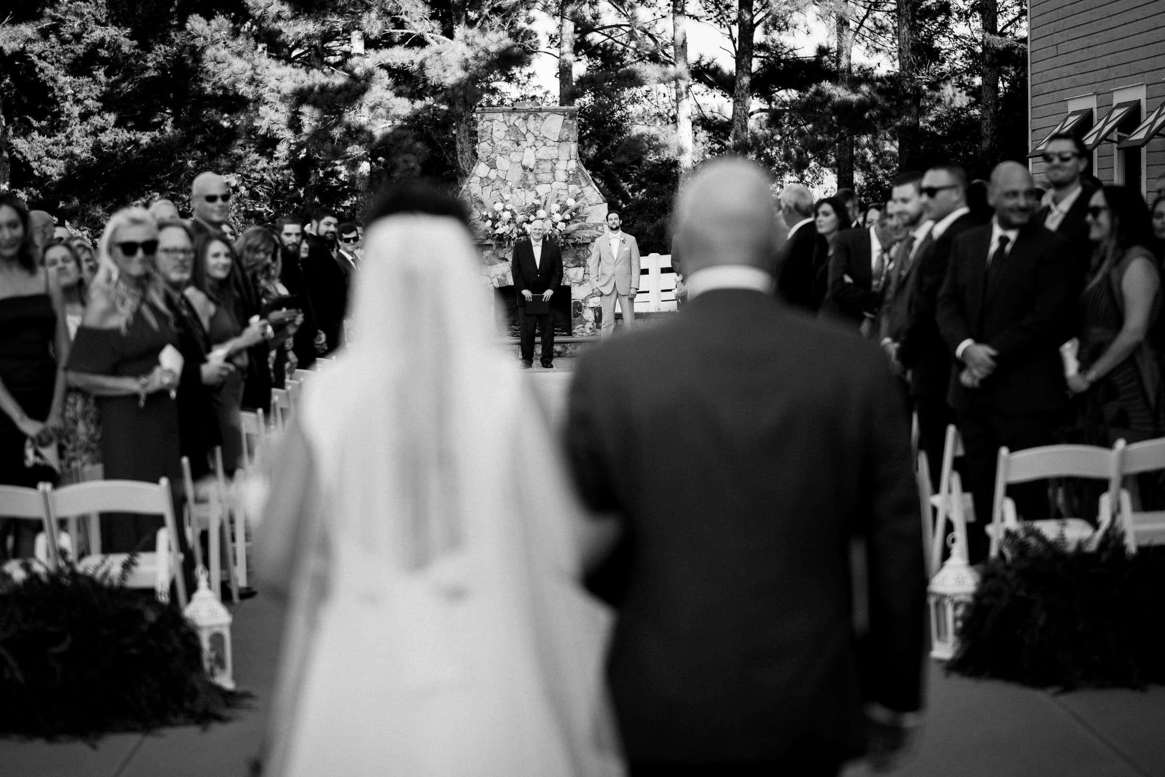 historic wakefield barn wedding - logan & rhett Black and white photo of a Wakefield Barn wedding ceremony. The bride and groom walk down the aisle lined with chairs and guests on both sides. The focus is on a figure at the altar ahead of them, with trees and a stone structure forming the backdrop.