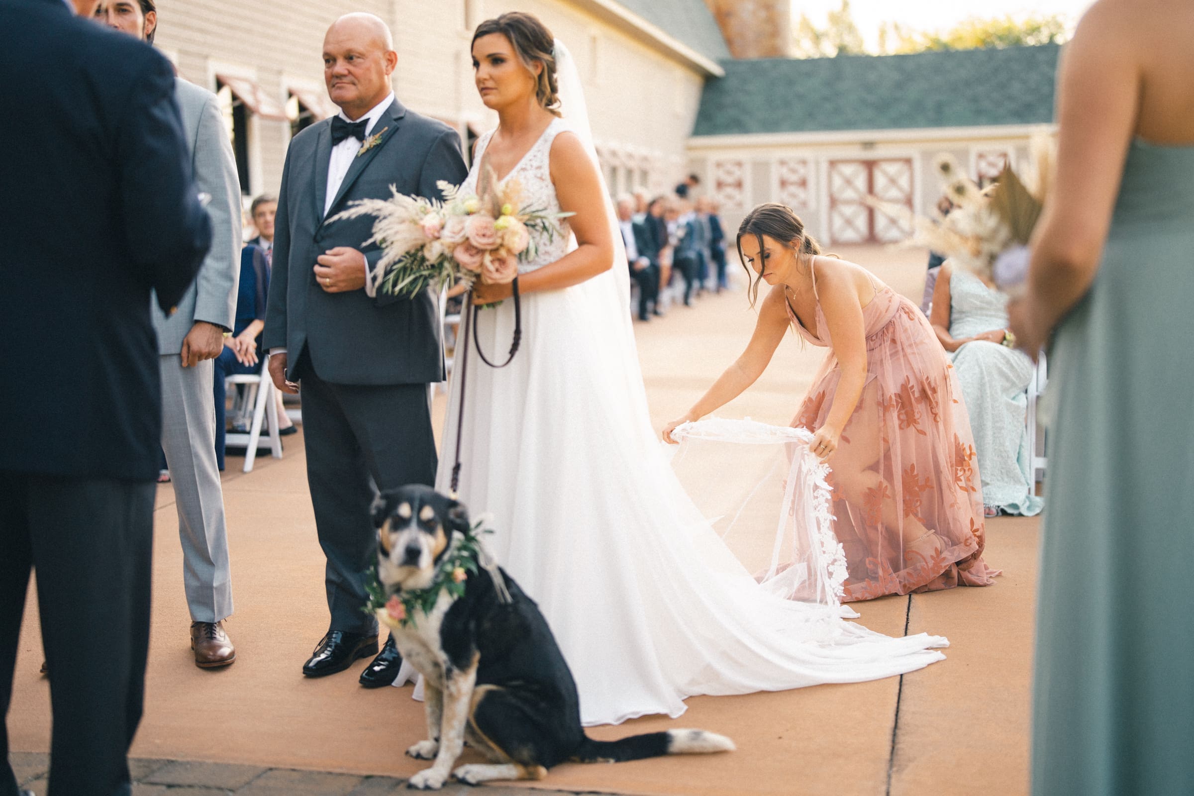 historic wakefield barn wedding - logan & rhett At a charming Wakefield Barn wedding, a bride in a white gown stands with the groom in a suit, holding a bouquet during their outdoor ceremony. A woman adjusts the bride's train, while a dog with a floral collar sits nearby and guests enjoy the picturesque scene.