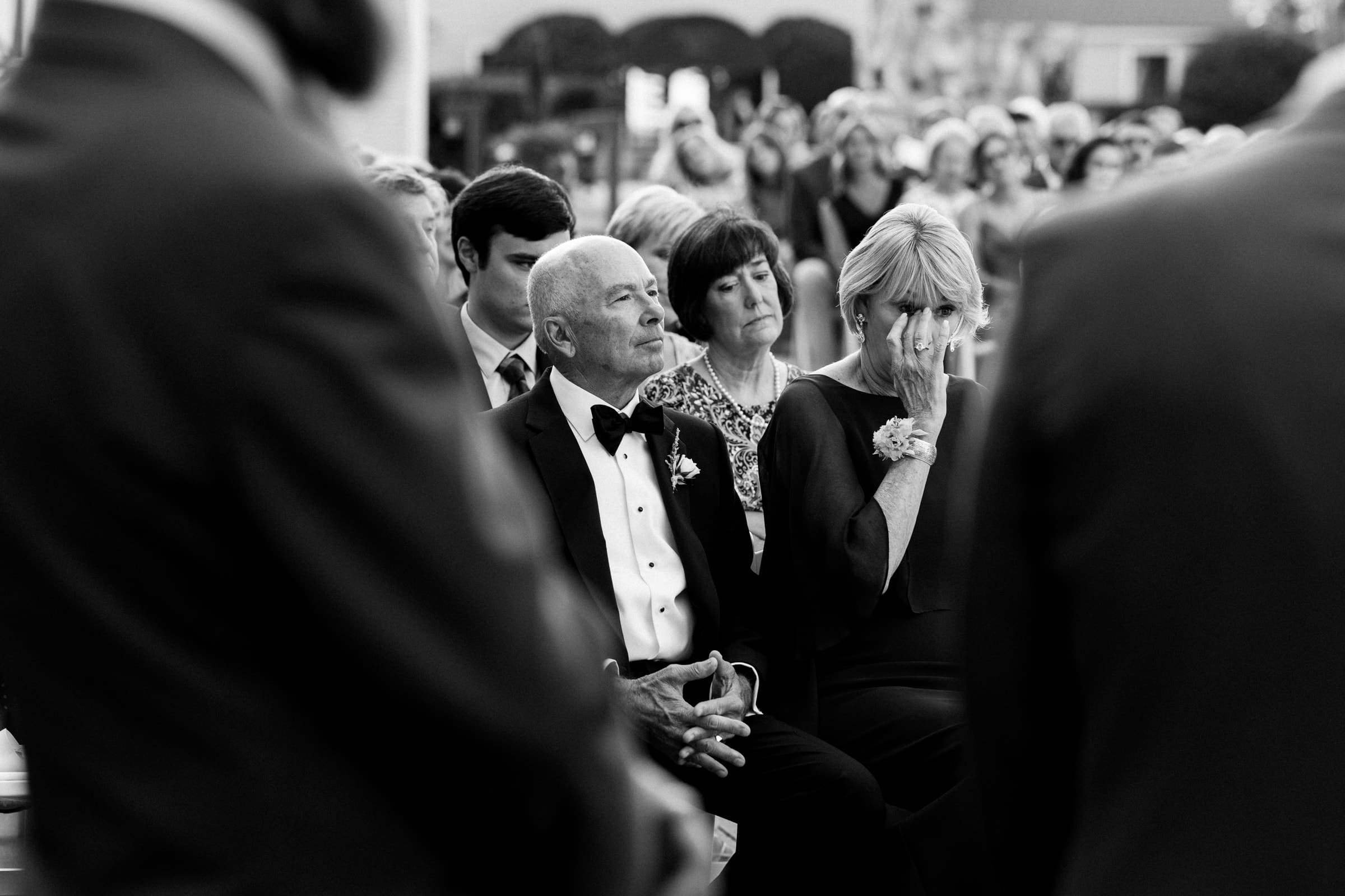 historic wakefield barn wedding - logan & rhett A black-and-white photo captures a Wakefield Barn wedding ceremony. An older, elegantly dressed couple sits in the front row, appearing emotional. The woman wipes tears while the man looks on thoughtfully. Blurred guests fill the background.