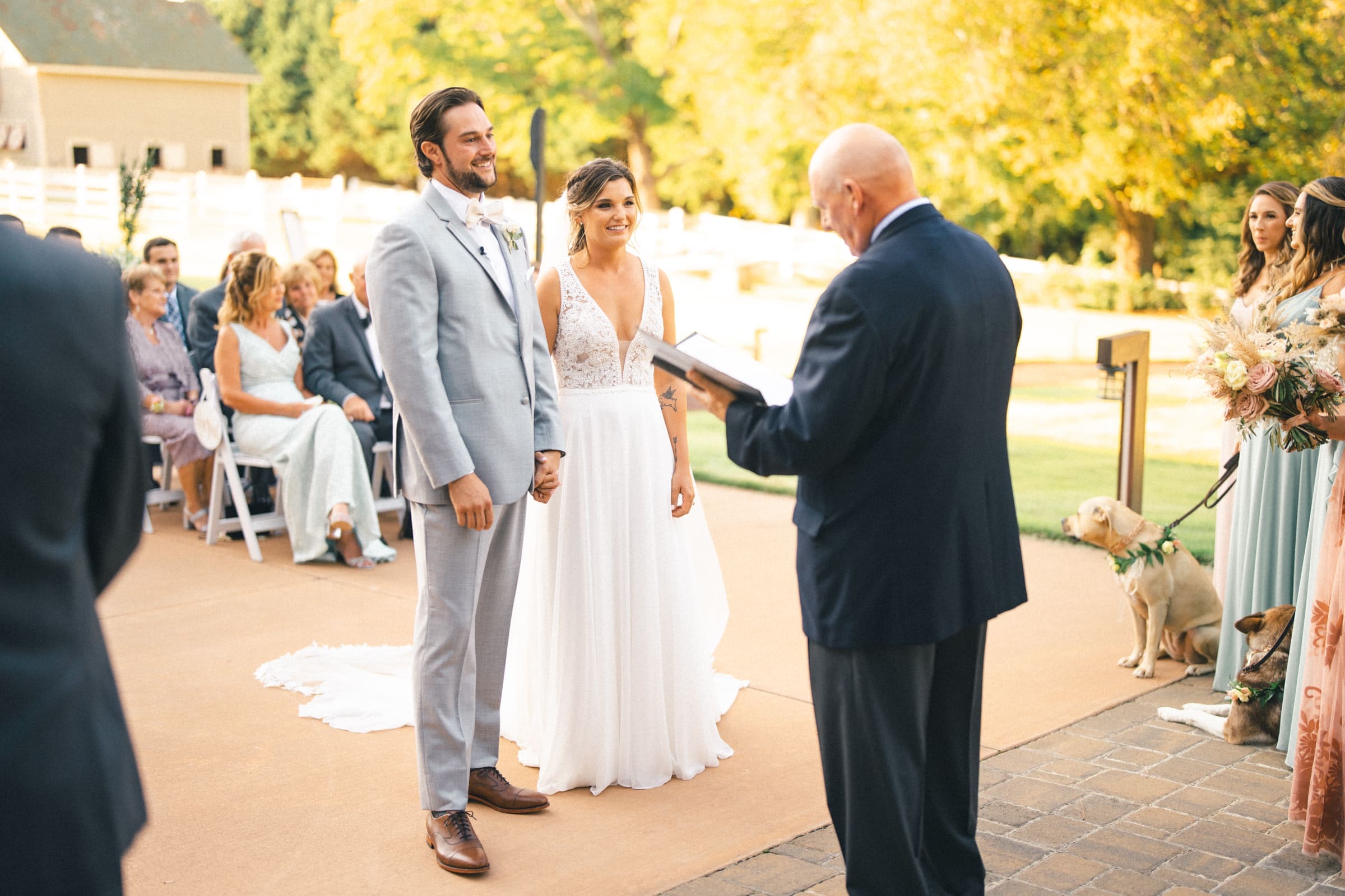 historic wakefield barn wedding - logan & rhett A bride and groom stand together outdoors during their Wakefield Barn wedding ceremony. The officiant reads from a book while wedding guests watch. The bridal party and a dog are visible to the side, set against trees and a sunny sky in the background.