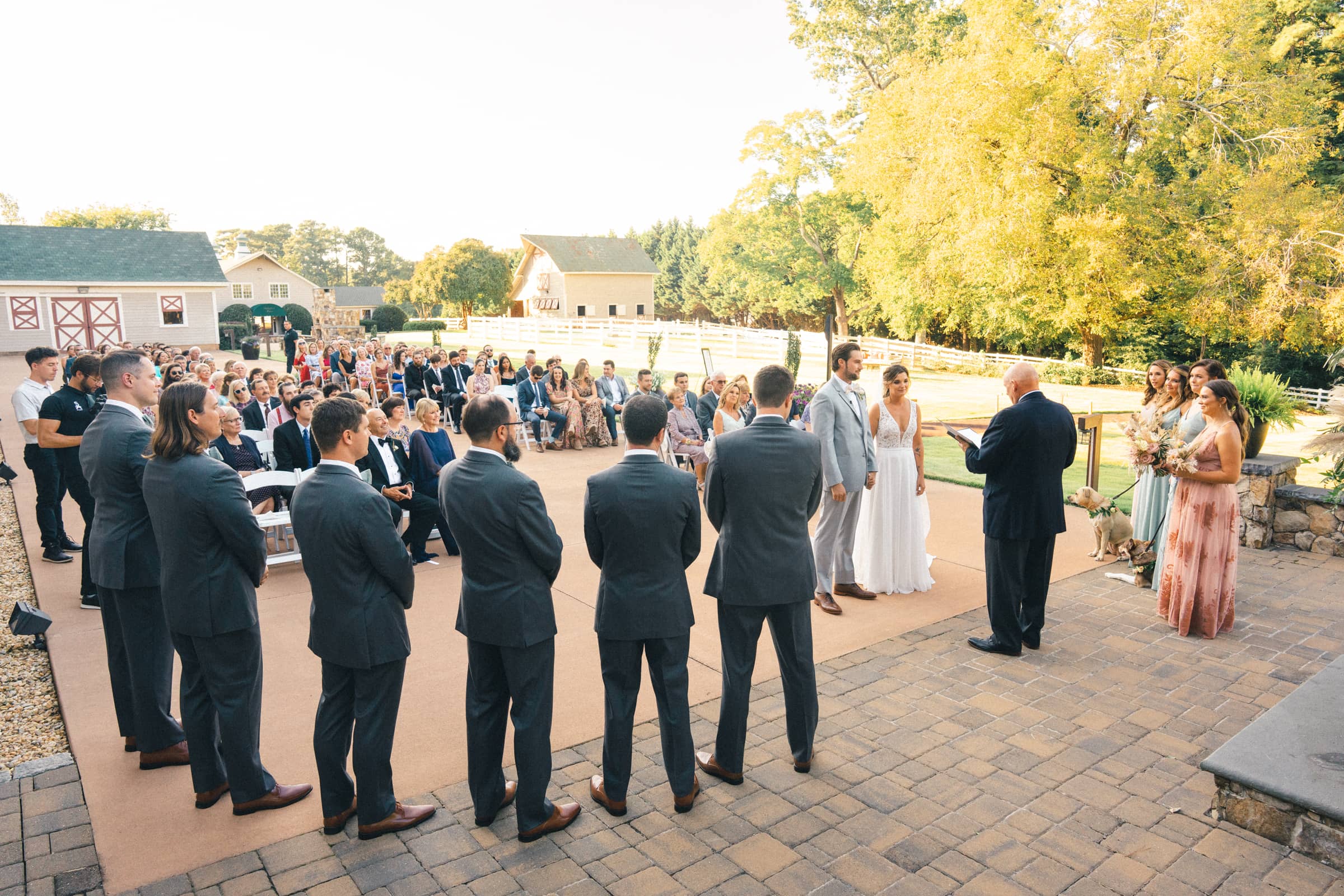 historic wakefield barn wedding - logan & rhett A Wakefield Barn wedding unfolds outdoors on a sunny day, where the couple stands facing the officiant, surrounded by bridesmaids and groomsmen. Guests are seated against a picturesque backdrop of trees and buildings, creating an enchanting atmosphere for this memorable celebration.