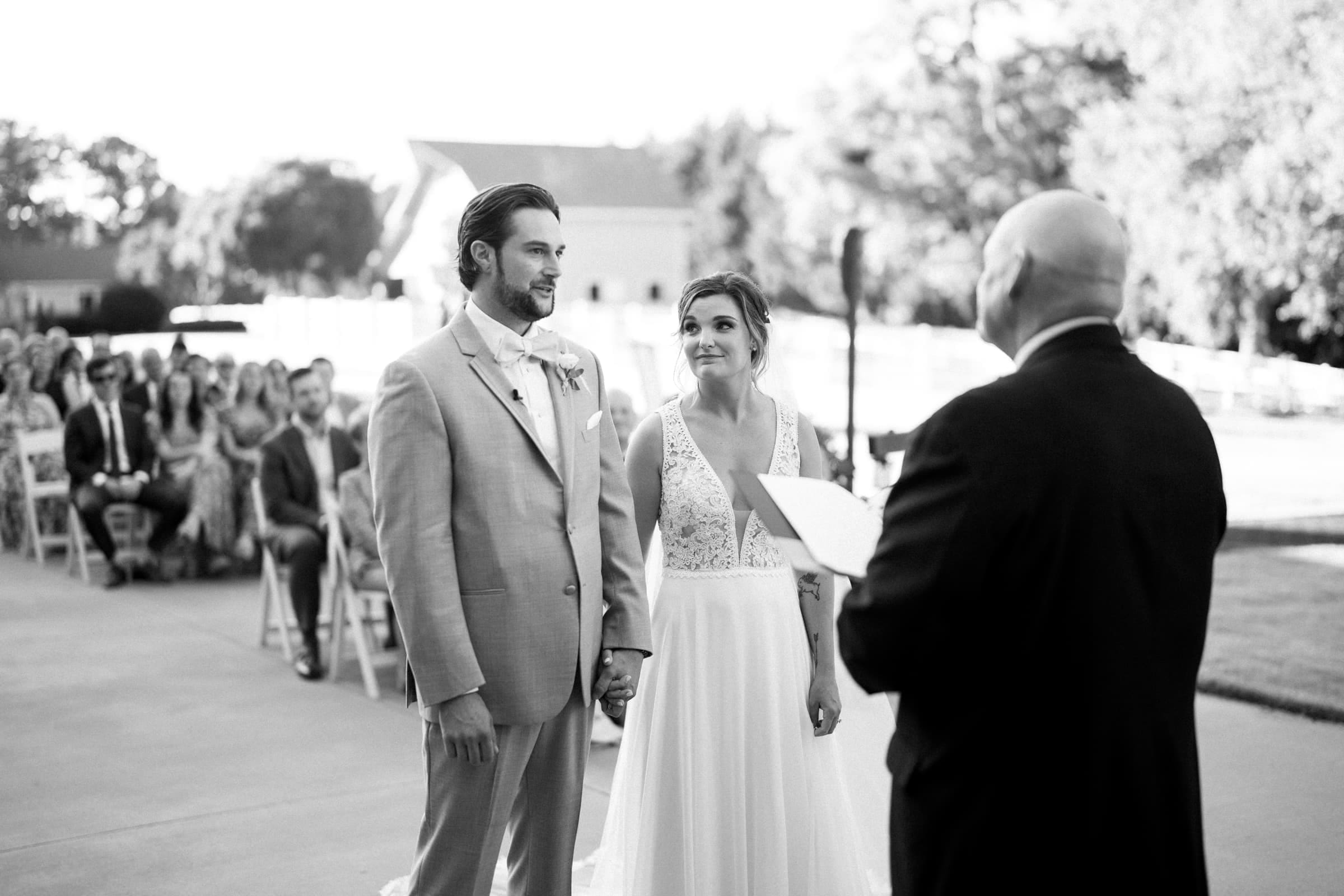 historic wakefield barn wedding - logan & rhett A black and white photo captures an outdoor wedding ceremony at Wakefield Barn. The couple holds hands, facing the officiant, while guests seated in rows watch the serene scene, with trees and buildings providing a picturesque backdrop.