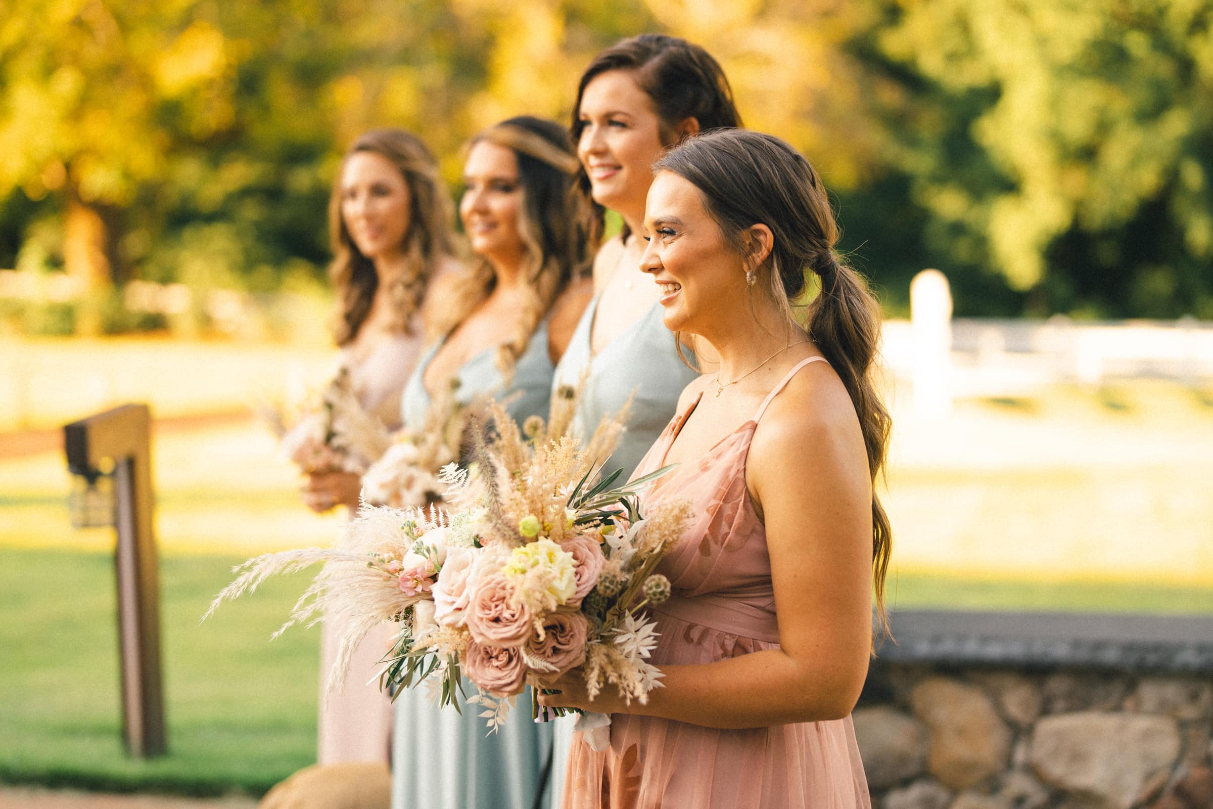 historic wakefield barn wedding - logan & rhett Four bridesmaids in pastel-colored dresses stand outdoors at a Wakefield Barn wedding, holding bouquets of flowers. They are smiling and looking in the same direction, with a sunny, green landscape in the background.