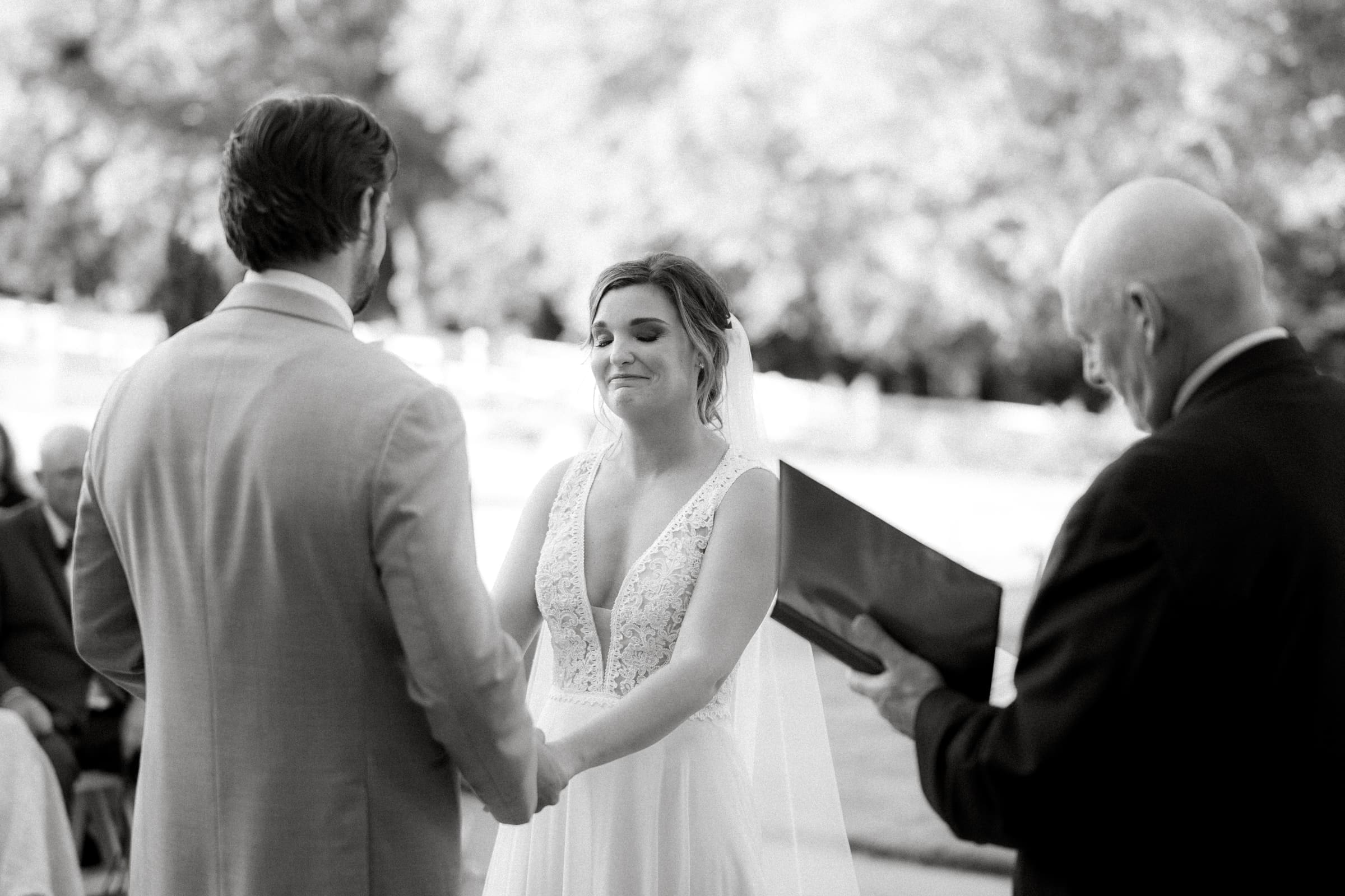 historic wakefield barn wedding - logan & rhett A bride and groom hold hands at their Wakefield Barn wedding, surrounded by softly blurred greenery. The bride dazzles in a white dress with lace details, while the groom complements her in a light suit. An officiant stands to the side, holding a book, ready to unite them both.