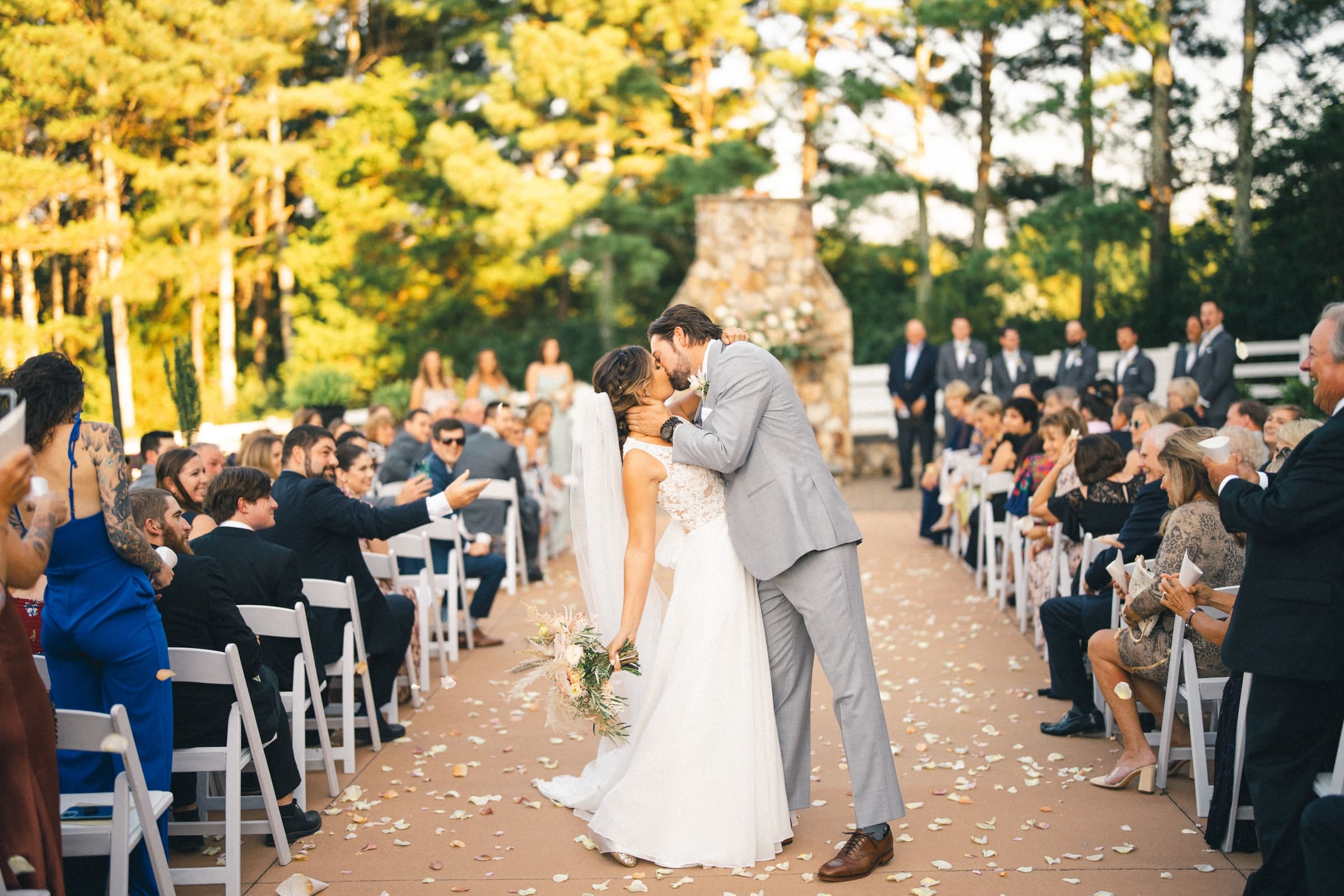 historic wakefield barn wedding - logan & rhett A bride and groom share a kiss at their Wakefield Barn wedding. They stand on a petal-strewn aisle surrounded by seated guests, with trees and a stone structure as the picturesque backdrop.