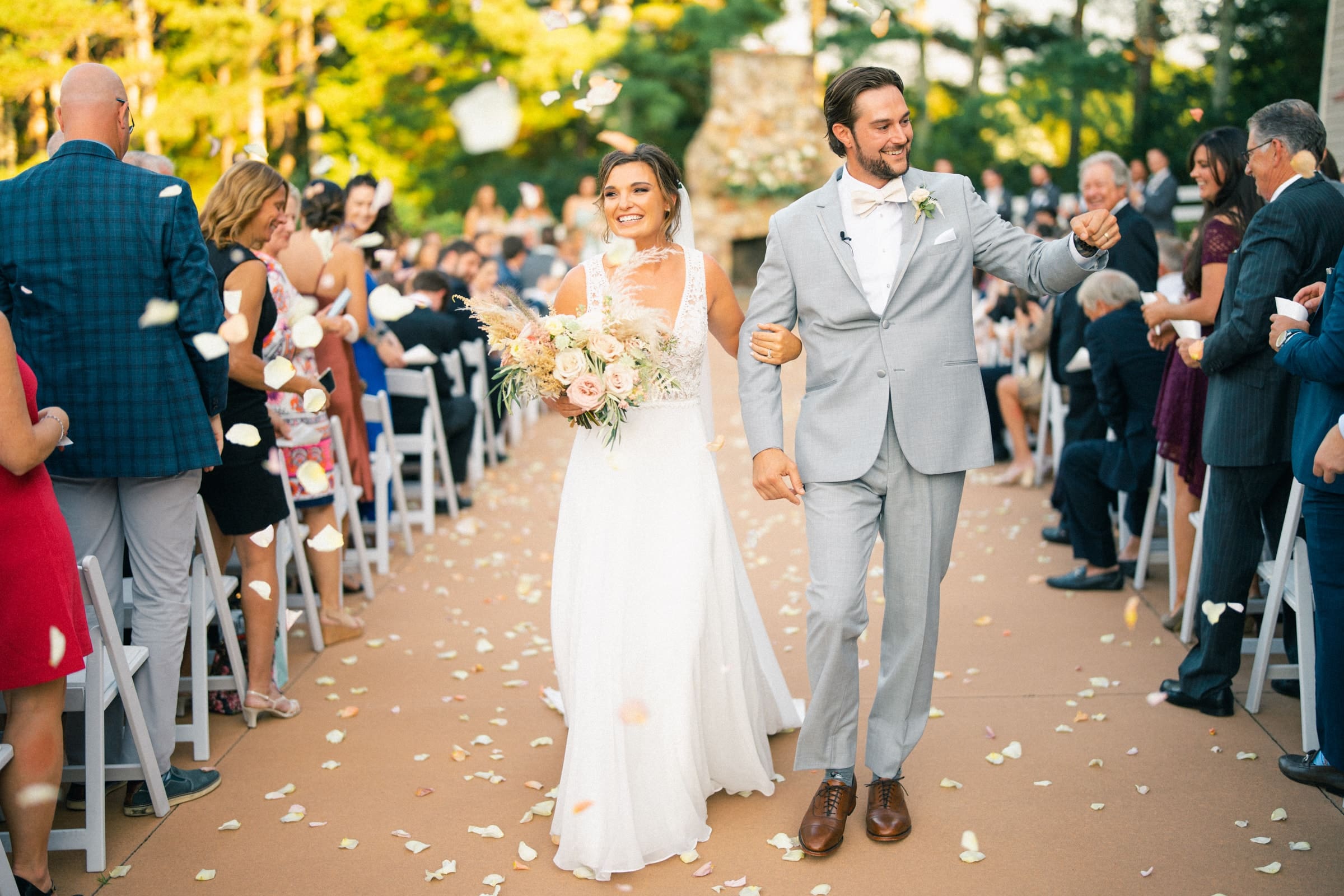 historic wakefield barn wedding - logan & rhett At the picturesque Wakefield Barn wedding, a newlywed couple walks down the aisle, smiling and holding hands. The bride, in a white dress with a bouquet, and the groom in a light gray suit bask under the sunny outdoor setting as guests on both sides shower them with flower petals.