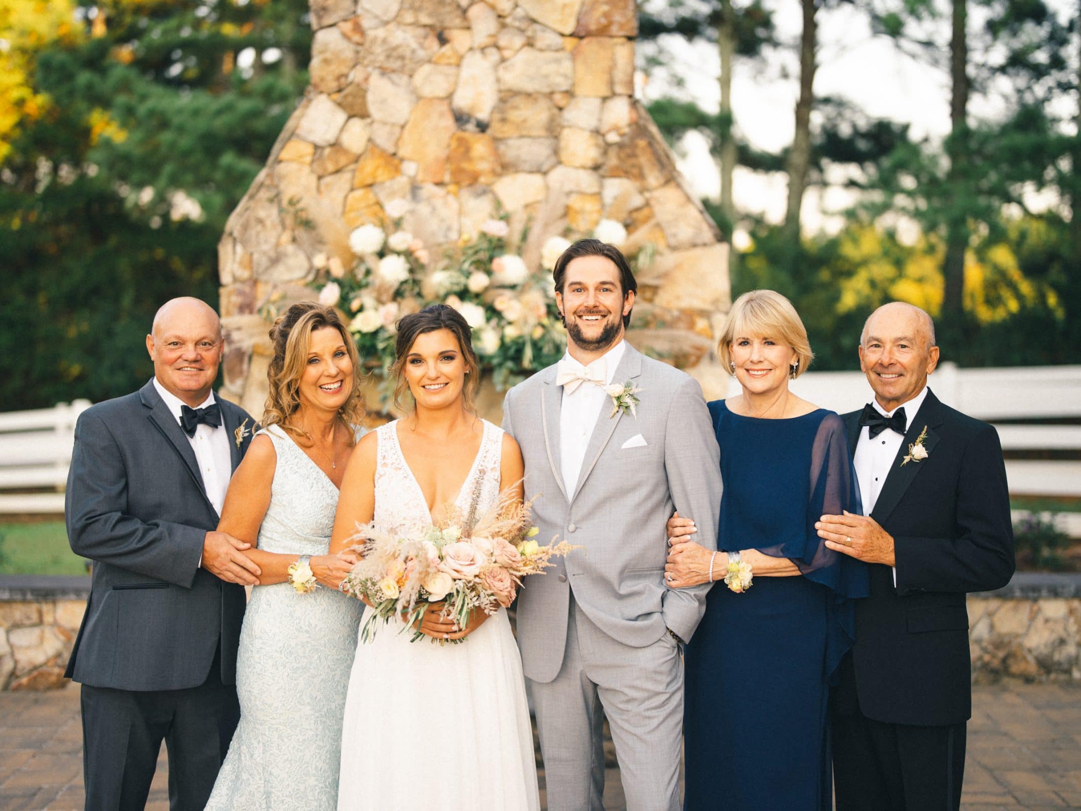 historic wakefield barn wedding - logan & rhett A bride and groom stand smiling in front of a stone structure at their Wakefield Barn wedding. Flanked by two older couples dressed in formal attire, they all pose cheerfully. The women hold bouquets, and a natural, wooded background completes the picturesque scene.
