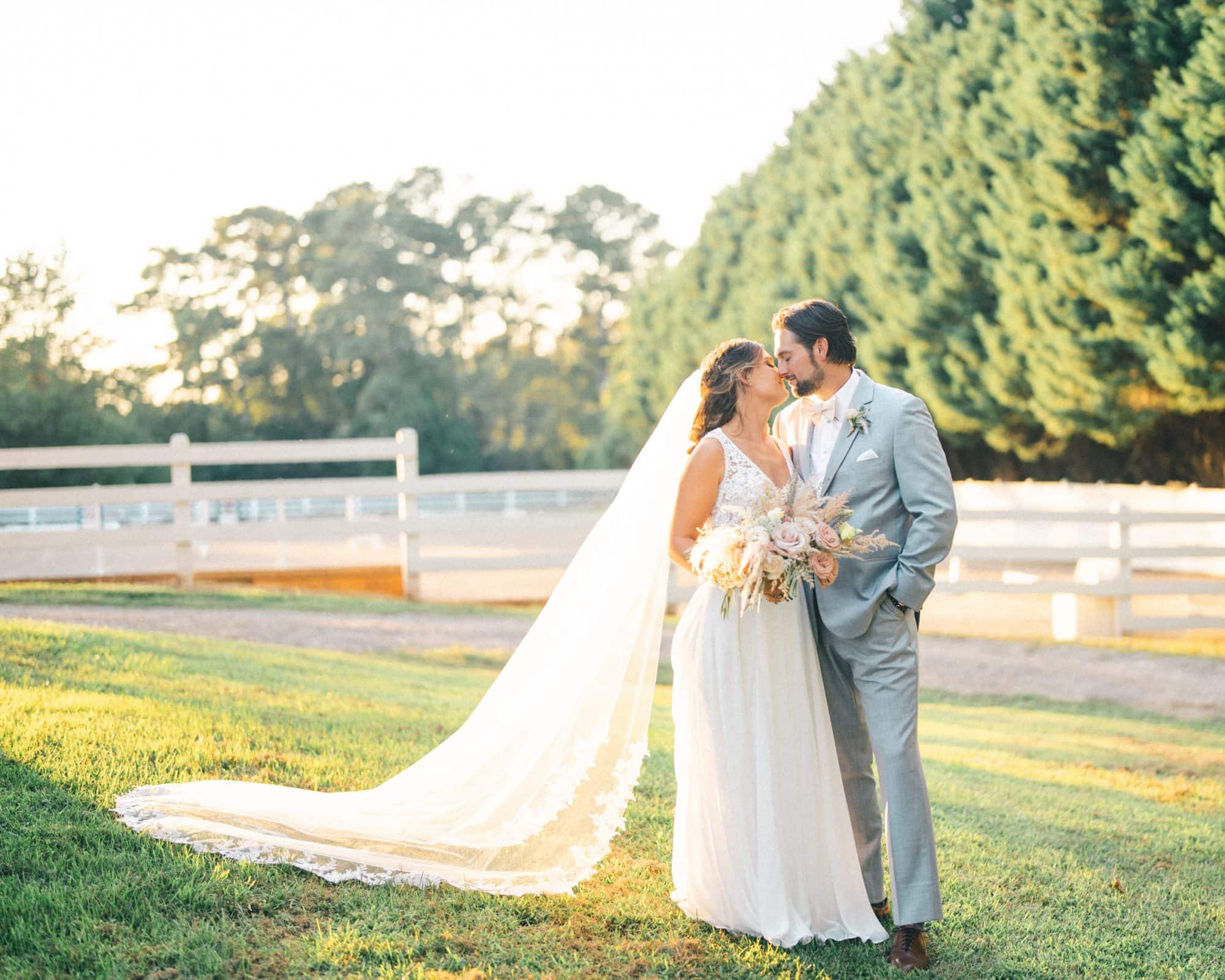 historic wakefield barn wedding - logan & rhett At their Wakefield Barn wedding, the bride and groom stand on a grassy area, gazing at each other lovingly. The bride holds a bouquet and wears a long veil, while the groom is in a light gray suit. A white fence and tall trees frame them under the bright sky.