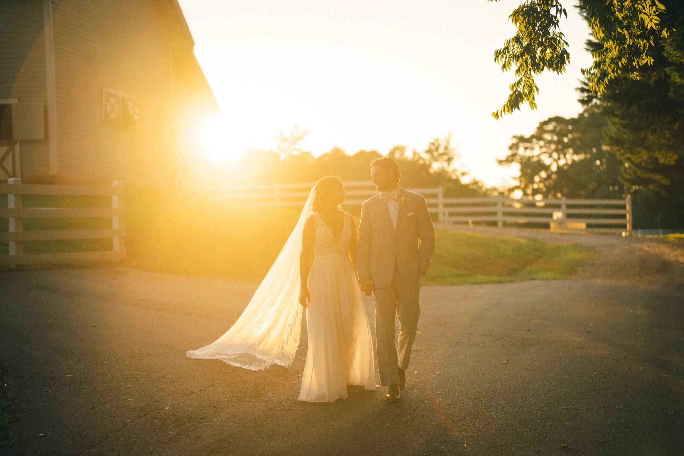 historic wakefield barn wedding - logan & rhett A bride and groom walk hand in hand on a sunlit path at Wakefield Barn. The bride wears a flowing white gown and veil, while the groom is in a light gray suit. The sun sets in the background, casting a warm glow over the enchanting wedding scene.