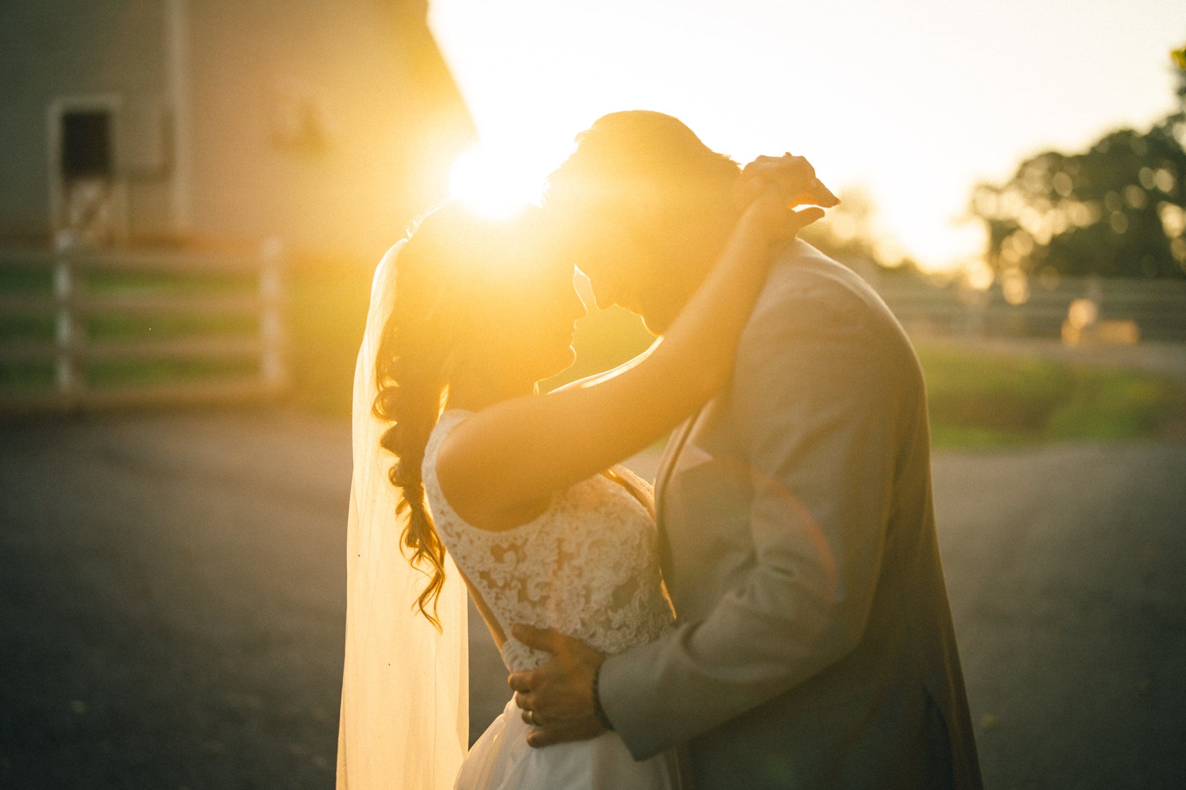 historic wakefield barn wedding - logan & rhett A bride and groom embrace in a romantic pose, silhouetted against a golden sunset at their Wakefield Barn wedding. The sun peeks through, creating a warm, glowing halo effect around them as they stand on a paved path with a farm-like setting in the background.