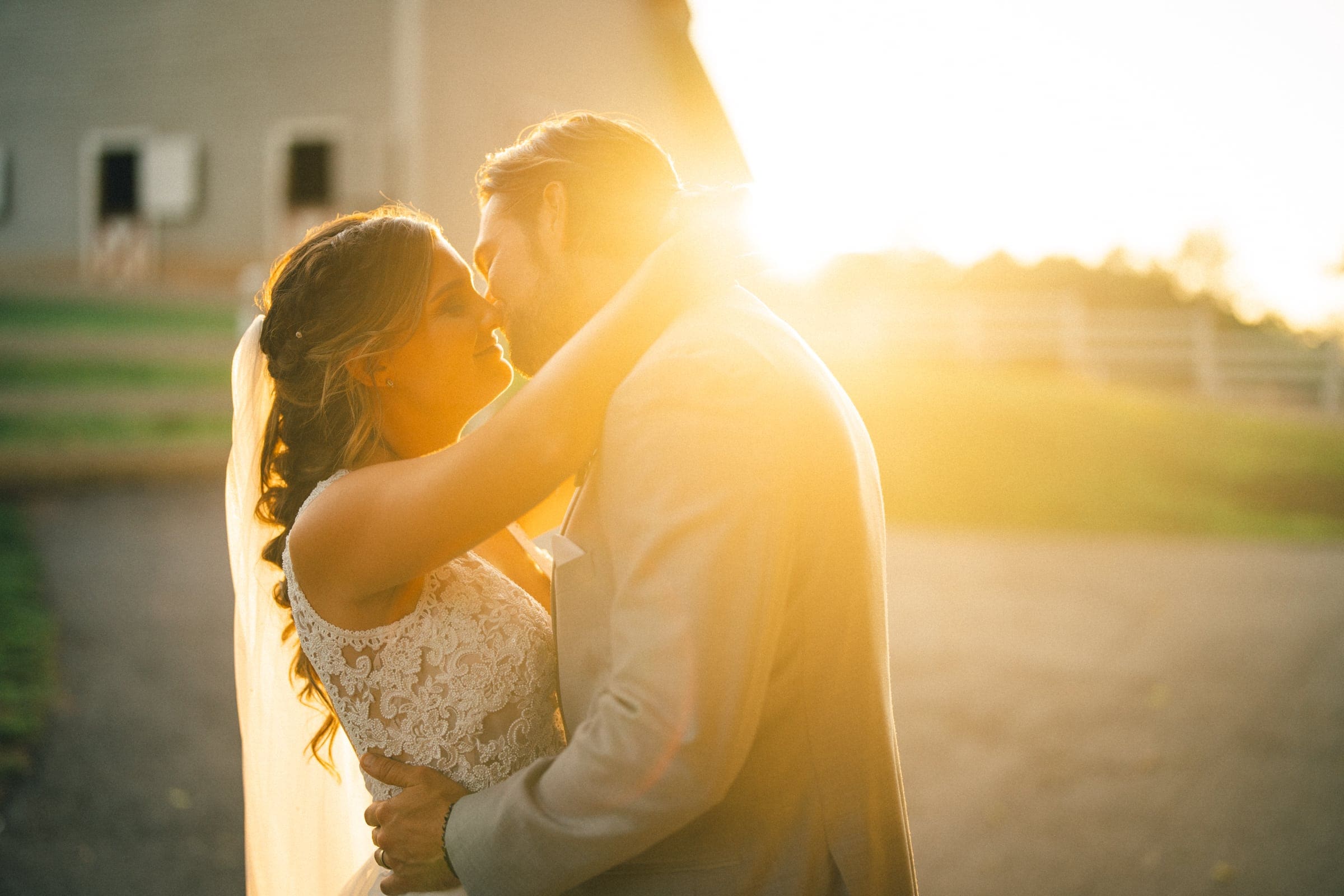 historic wakefield barn wedding - logan & rhett A couple embraces and kisses at sunset, surrounded by a warm golden glow at their Wakefield Barn wedding. The woman wears a white lace dress with a veil, and the man is in a light-colored suit. A barn and fence are visible in the blurred background.