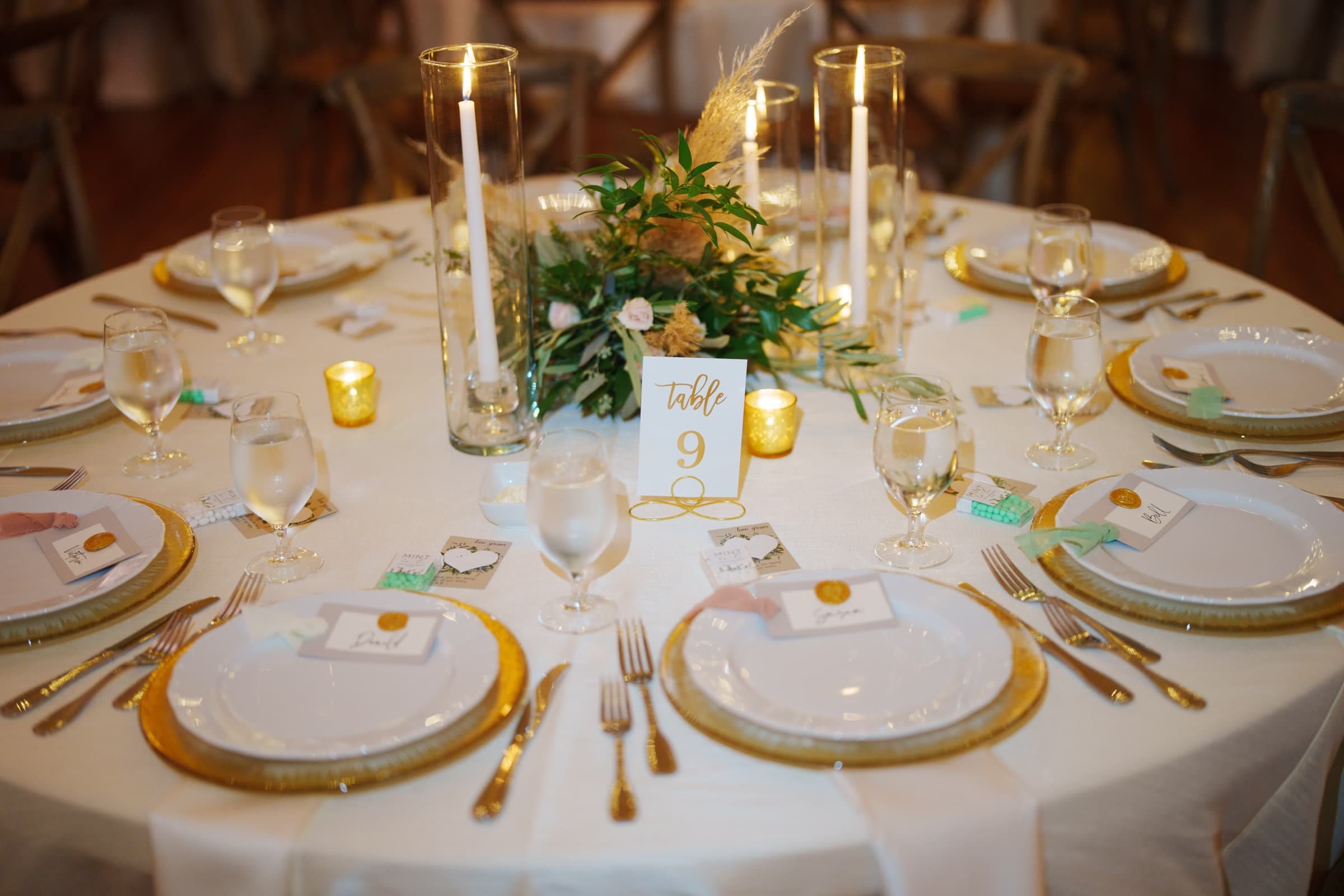 historic wakefield barn wedding - logan & rhett A beautifully set round table for a formal Wakefield Barn wedding, featuring a white tablecloth, gold-trimmed plates, tall candles, and a lush green centerpiece. A card in the center reads Table 9. Glasses and cutlery are neatly arranged for each guest.