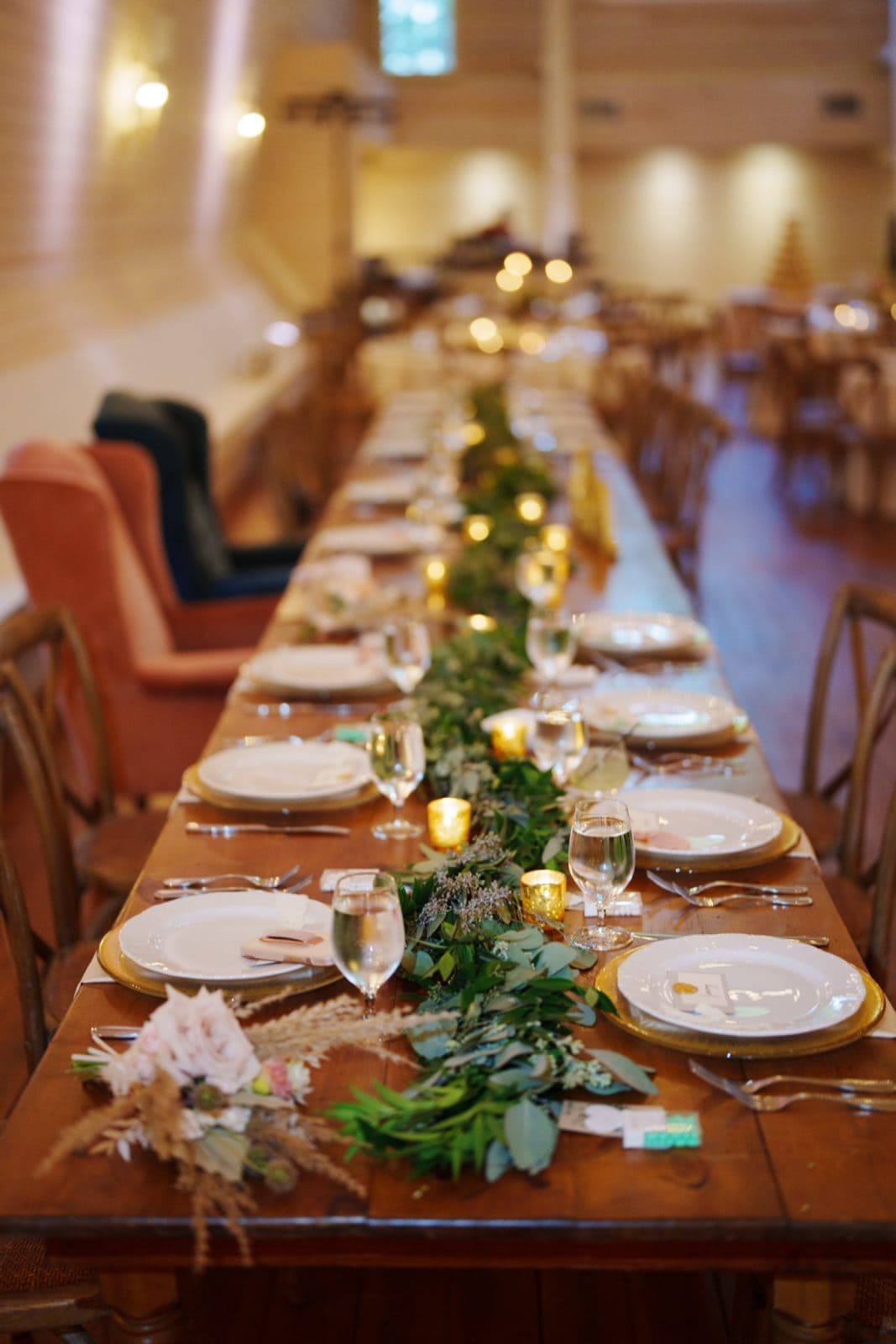 historic wakefield barn wedding - logan & rhett A long wooden dining table is elegantly set with white plates, wine glasses, and floral centerpieces at the Wakefield Barn wedding. Greenery and small candles are arranged along the center, while soft lighting in the background creates a warm, inviting atmosphere.