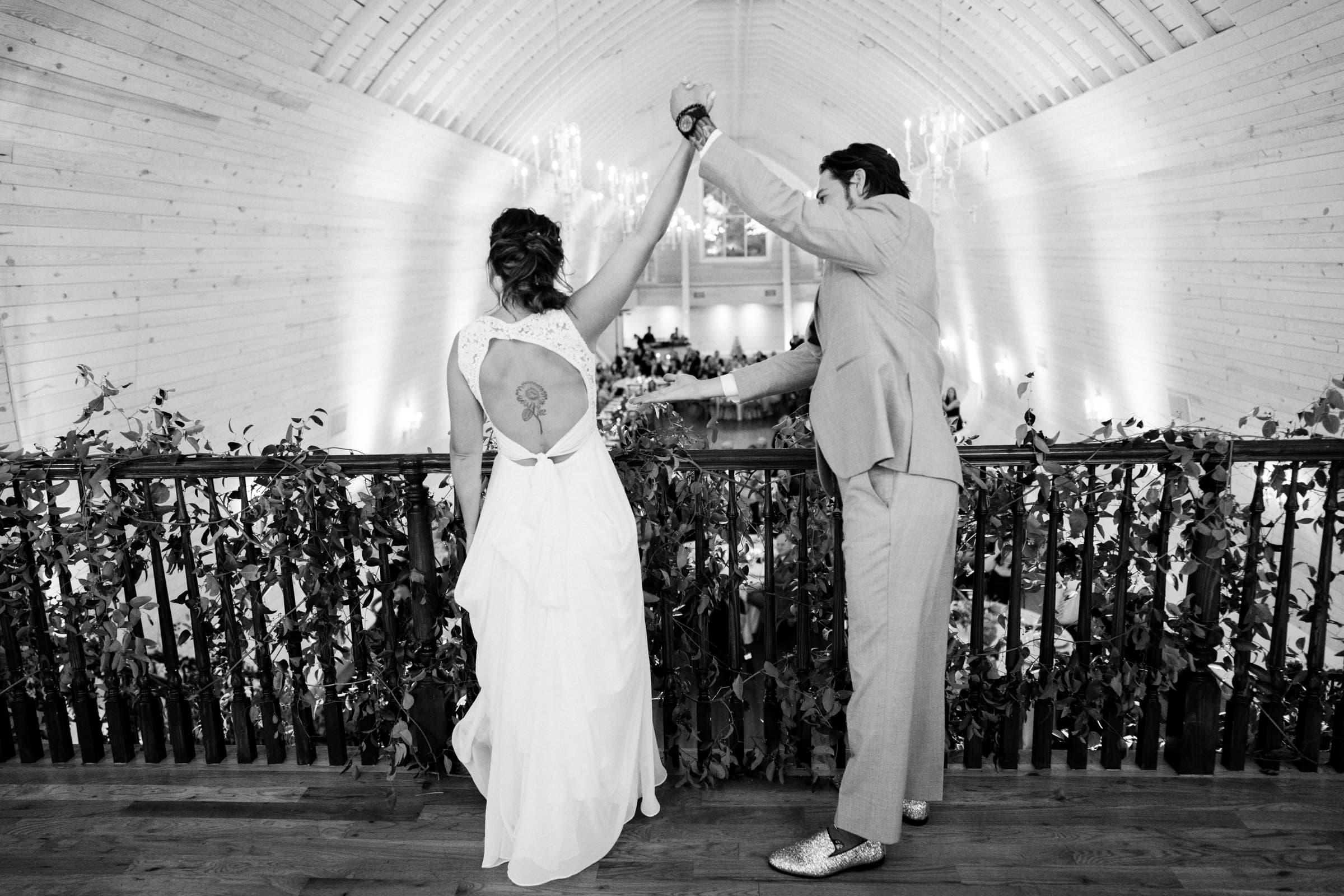 historic wakefield barn wedding - logan & rhett A couple in formal attire stands on the balcony at Wakefield Barn, facing away from the camera. They hold hands as the woman's dress, with its open back revealing a tattoo, catches the eye. The venue, elegantly illuminated and adorned with greenery and chandeliers, exudes charm.
