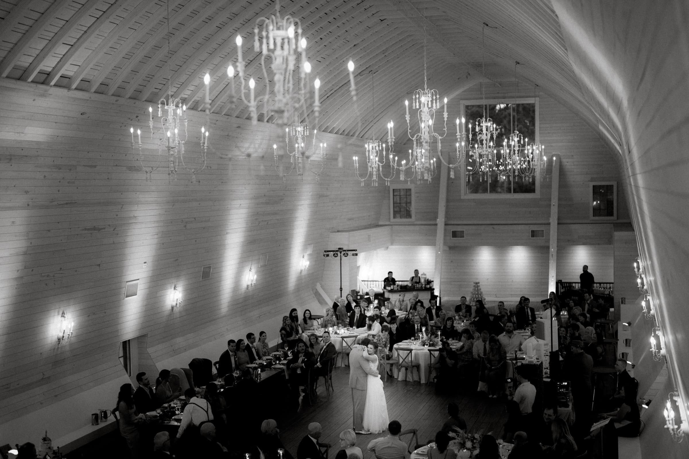 historic wakefield barn wedding - logan & rhett A black-and-white photo of a Wakefield Barn wedding reception in a large, elegant hall with chandeliers. A couple is dancing at the center while guests sit at tables watching. The room boasts high, vaulted ceilings and wooden walls.