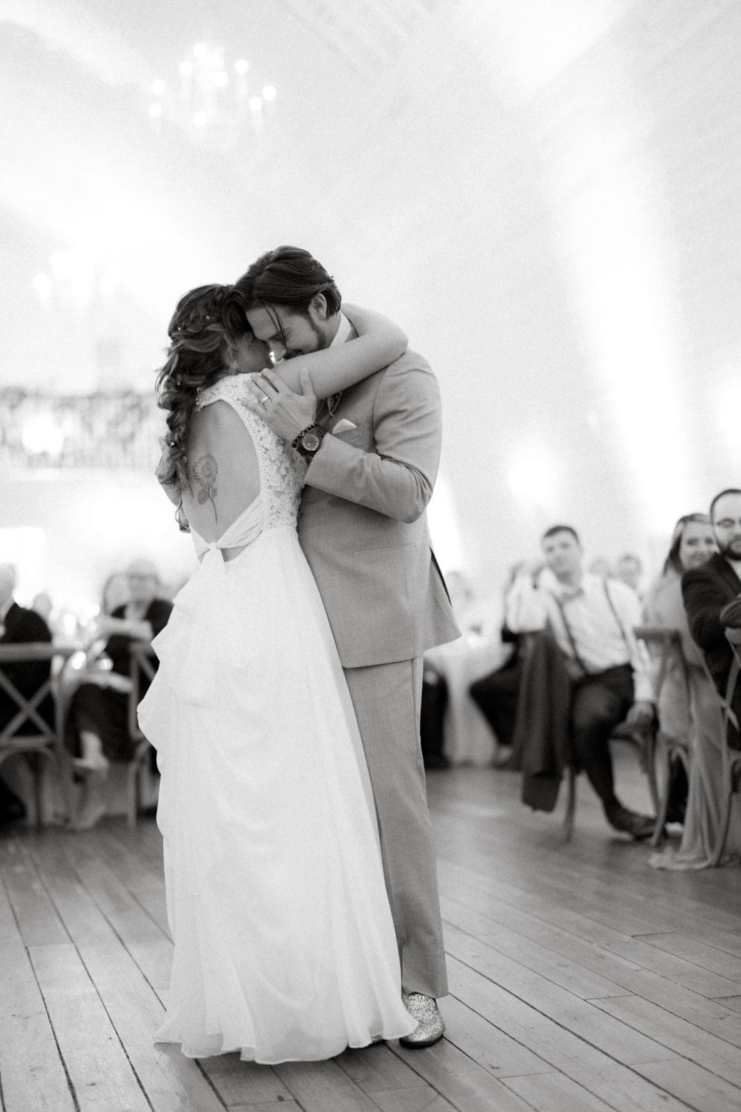 historic wakefield barn wedding - logan & rhett At the Wakefield Barn wedding, a bride and groom share a tender dance at their reception. They are in an elegantly lit venue with guests seated in the background. The bride wears a flowing white gown, while the groom is in a light-colored suit.