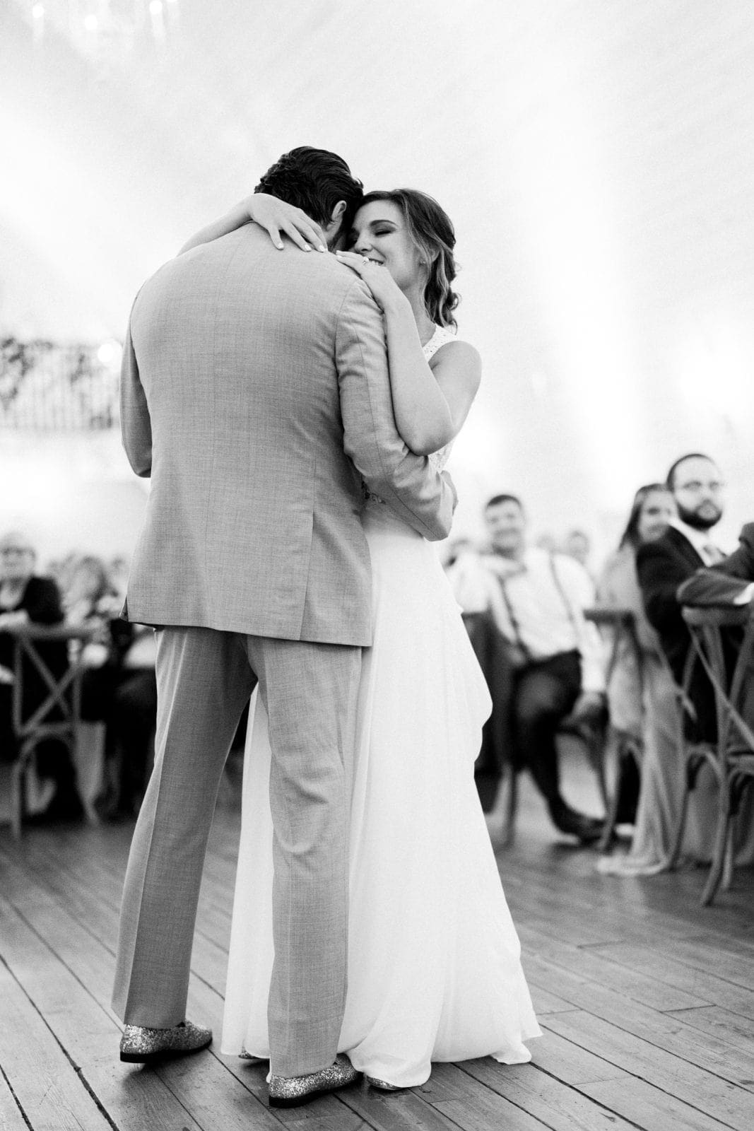 historic wakefield barn wedding - logan & rhett A couple dances closely at the Wakefield Barn wedding, surrounded by seated guests in a warmly lit venue. The groom wears a light suit, and the bride is in a white gown. The scene is intimate and joyful, capturing a tender wedding moment.