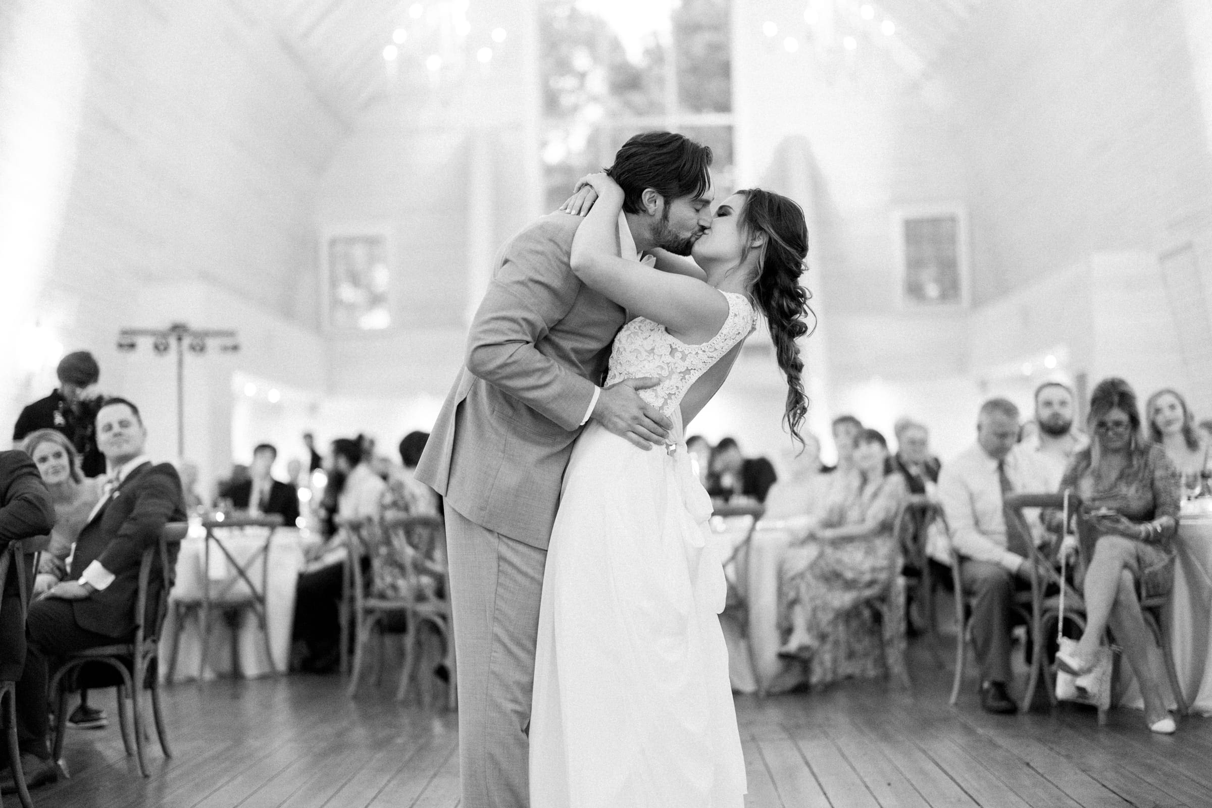 historic wakefield barn wedding - logan & rhett At the Wakefield Barn wedding, a bride and groom share a romantic first dance in a softly lit reception hall. Guests seated at tables watch the couple. The black and white image captures the elegance and joy of the moment.