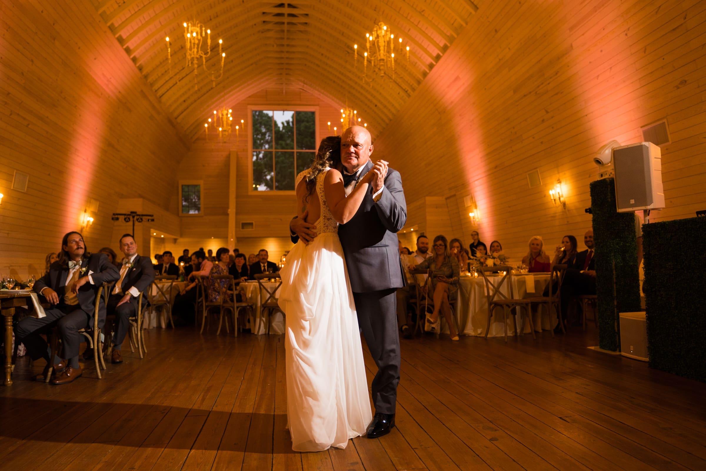 historic wakefield barn wedding - logan & rhett A bride and groom share their first dance in the warmly lit Wakefield Barn venue. Guests are seated at tables in the background, watching and smiling. Chandelier lights hang from the wooden ceiling, adding a romantic ambiance to this enchanting Wakefield Barn wedding scene.