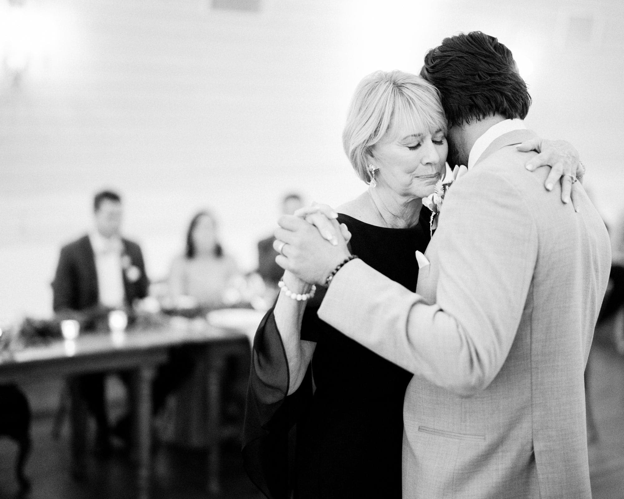 historic wakefield barn wedding - logan & rhett At a Wakefield Barn wedding, a woman and a man share a heartfelt dance. The grayscale image captures their embrace on the dimly lit dance floor, with seated guests blurred in the background, evoking an intimate and emotional atmosphere.