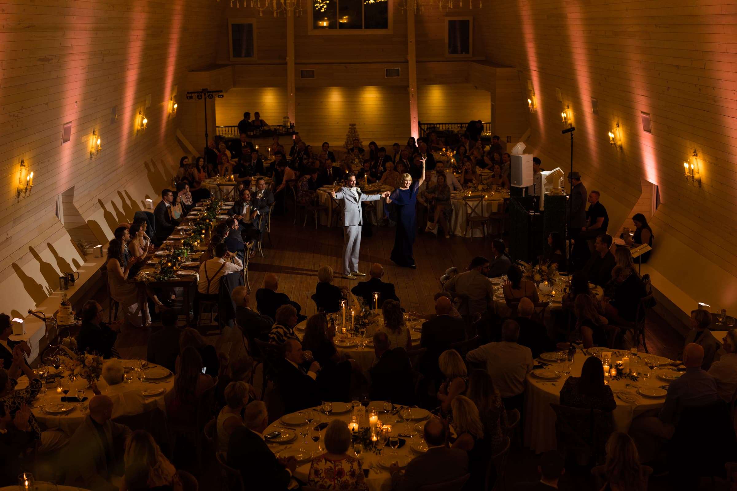 historic wakefield barn wedding - logan & rhett A dimly lit, elegant Wakefield Barn wedding reception unfolds inside a wooden hall. Guests seated at round tables with candles watch a couple dance in the center of the room. The walls are adorned with warm lighting, creating a cozy atmosphere.