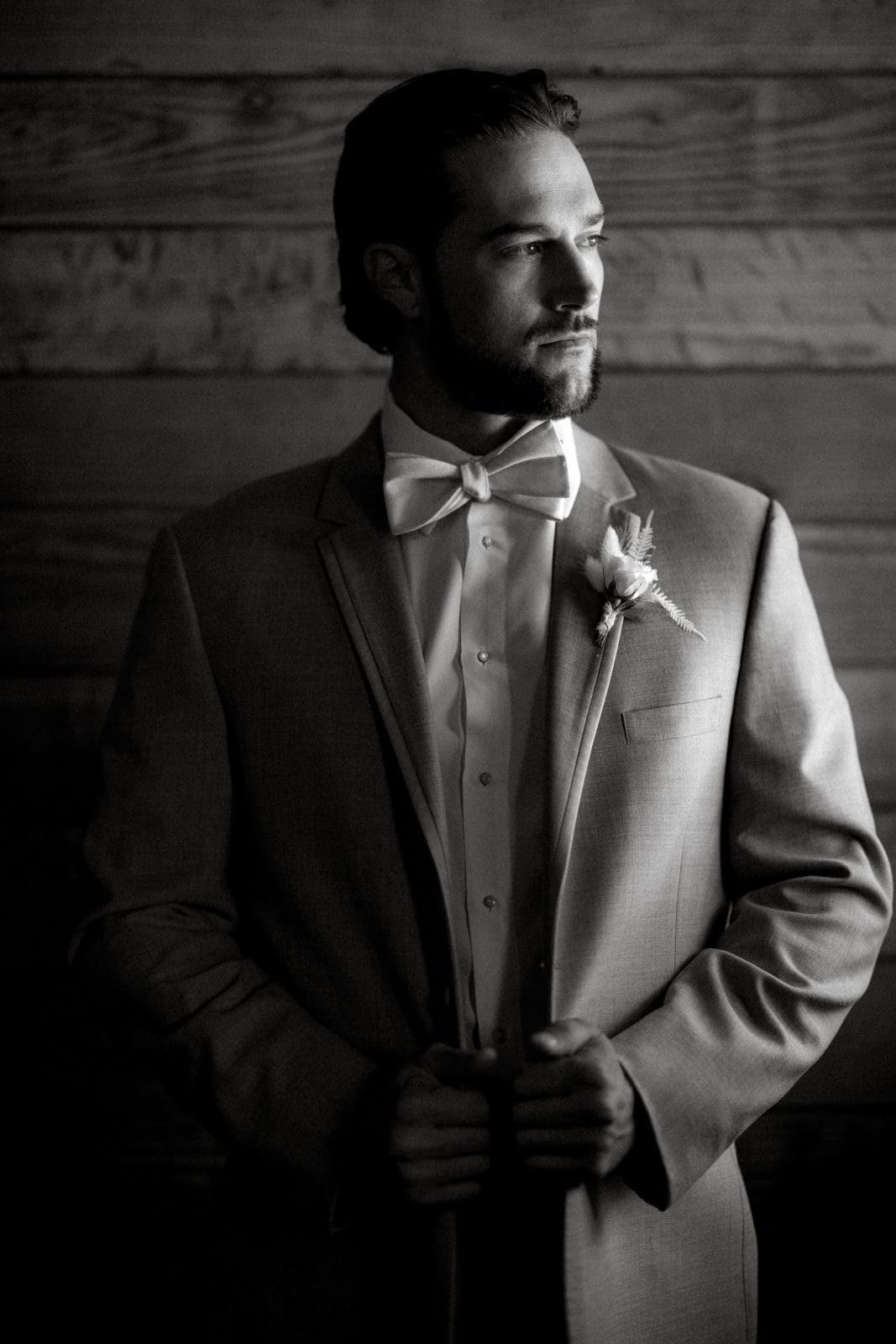 historic wakefield barn wedding - logan & rhett Black and white photo of a man in a suit and bow tie, looking thoughtful with hands clasped, standing at the Wakefield Barn wedding. A boutonniere is pinned to his jacket against a backdrop of wooden planks.