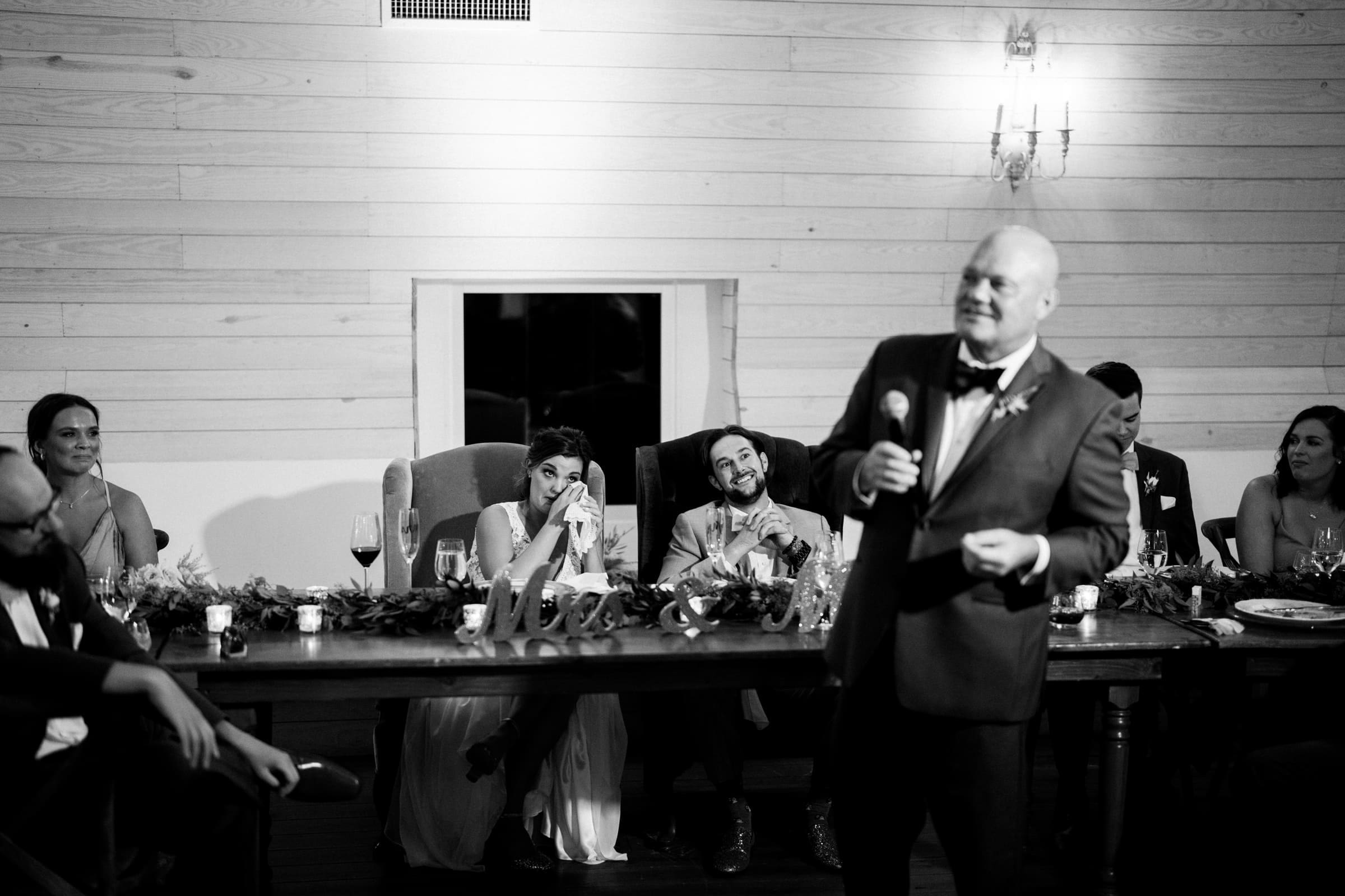 historic wakefield barn wedding - logan & rhett A black and white photo captures a Wakefield Barn wedding reception. A man in a suit delivers a heartfelt speech as the bride, seated at a long table, smiles and covers her face with her hand. Nearby, the groom and other guests listen intently to the moment.