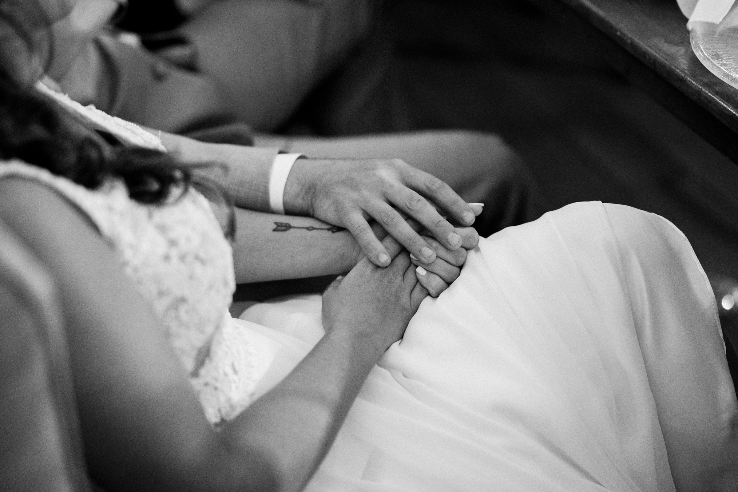 historic wakefield barn wedding - logan & rhett A black and white photo captures a couple closely seated, holding hands at their Wakefield Barn wedding. The woman in a lacy dress and the man in a suit share an intimate moment, with the focus on their connected hands highlighted by a tattoo on the woman's forearm.