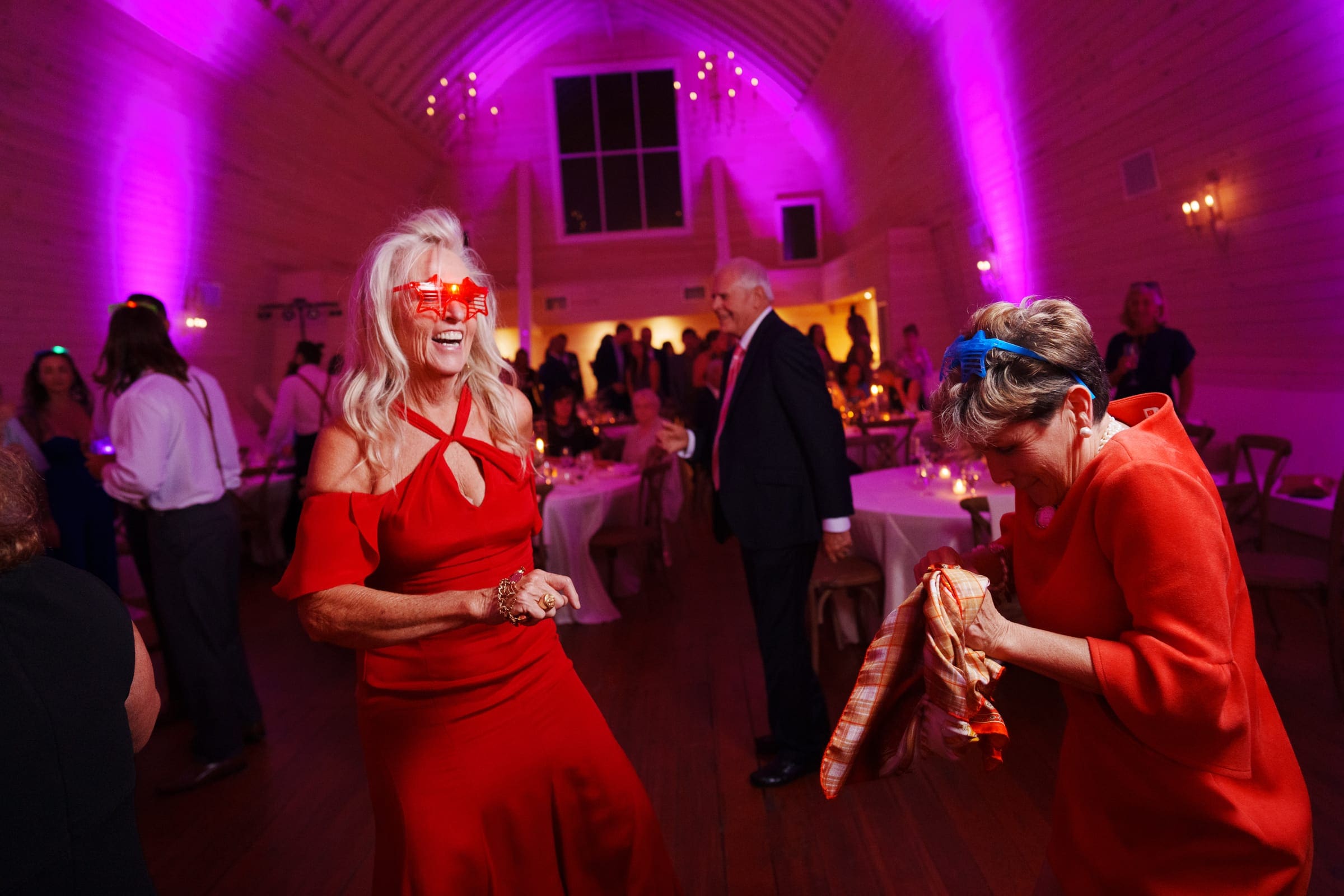 historic wakefield barn wedding - logan & rhett Two women in red dresses dance joyfully at the lively Wakefield Barn wedding, wearing novelty glasses. The venue is illuminated with purple lights, creating a festive atmosphere. Guests in the background enjoy the celebration in a spacious, elegantly lit hall.