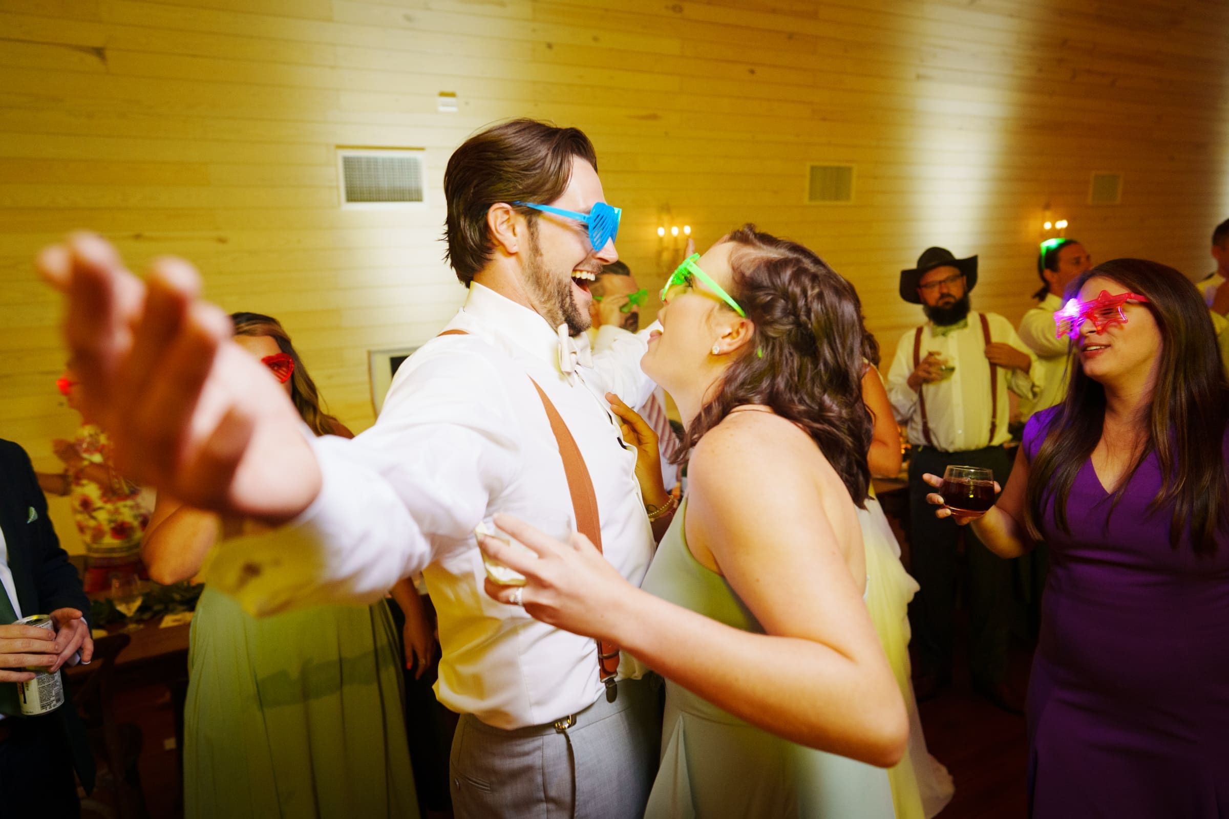 historic wakefield barn wedding - logan & rhett At a lively indoor party reminiscent of a Wakefield Barn wedding, people are dancing and laughing, donning colorful sunglasses and festive clothing. The room's warm wooden interior adds to the charm as guests in the background savor drinks, fully immersed in celebration.
