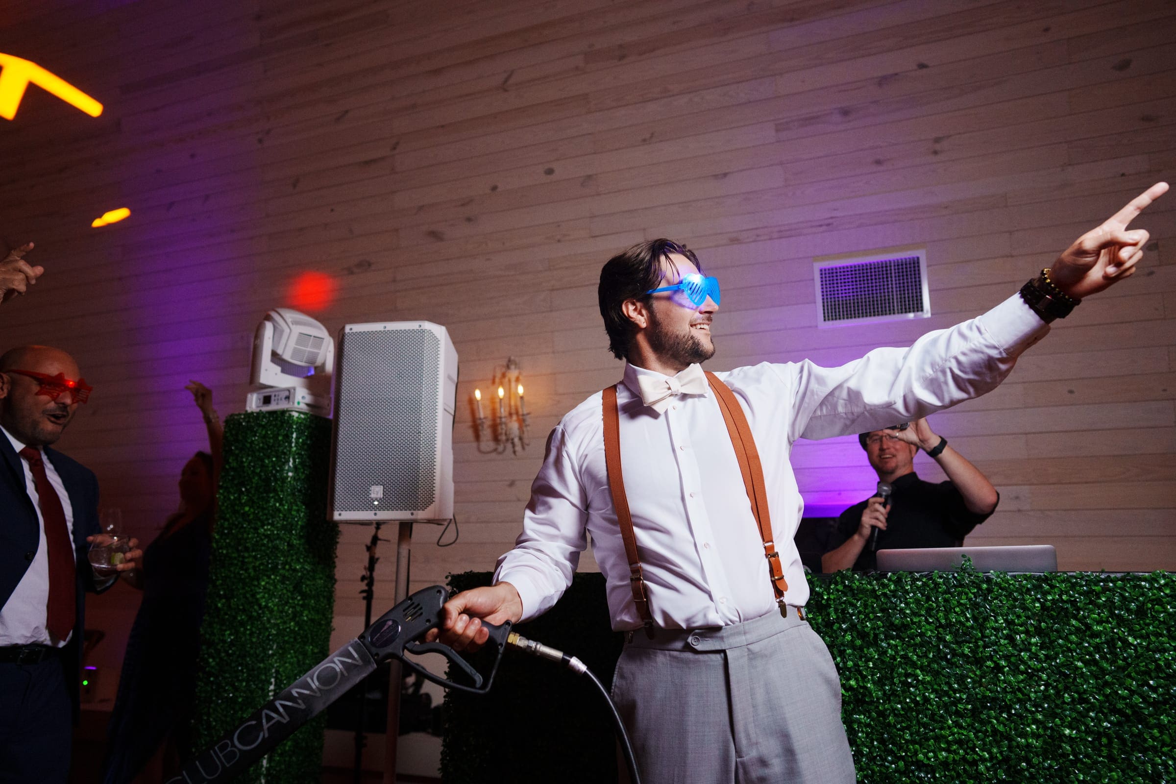 historic wakefield barn wedding - logan & rhett At a lively Wakefield Barn wedding, a man in a white shirt, bow tie, and suspenders dances energetically while wielding a CO2 cannon. He's sporting glowing glasses as colorful lights illuminate the wooden walls. A DJ spins tracks behind him, adding to the festive atmosphere.