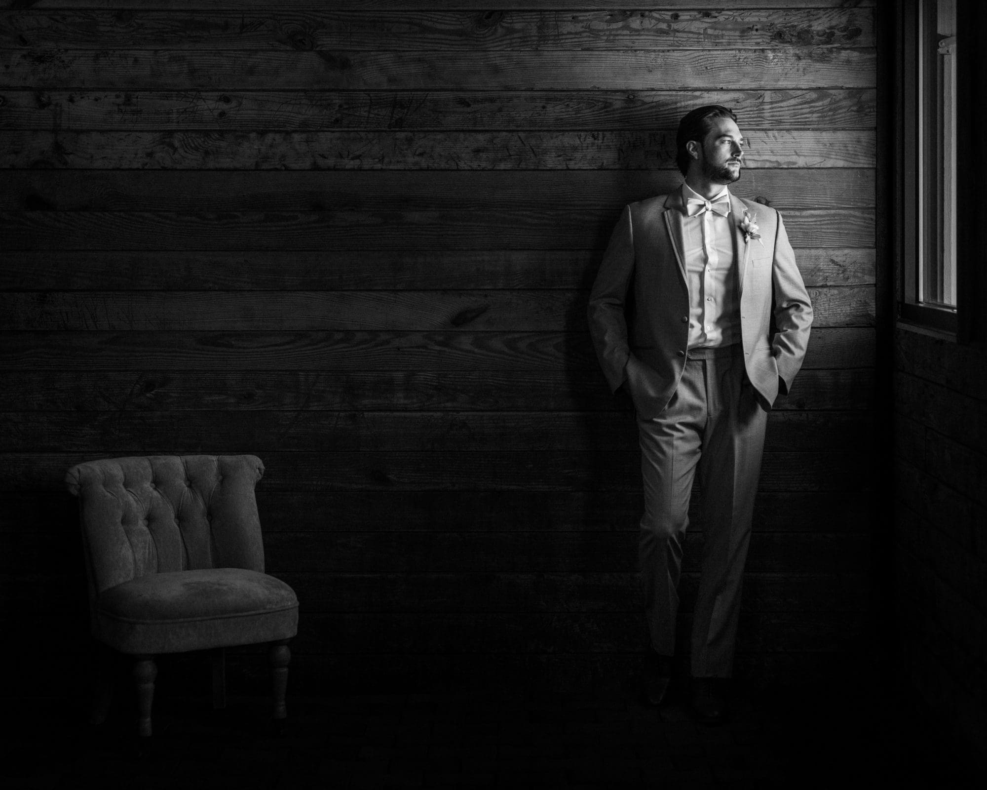 A man in a suit and bow tie stands in a dimly lit room at Wakefield Barn, leaning against a wooden wall and gazing out a window. A tufted chair is positioned nearby. The scene, captured in black and white, exudes a contemplative wedding atmosphere.