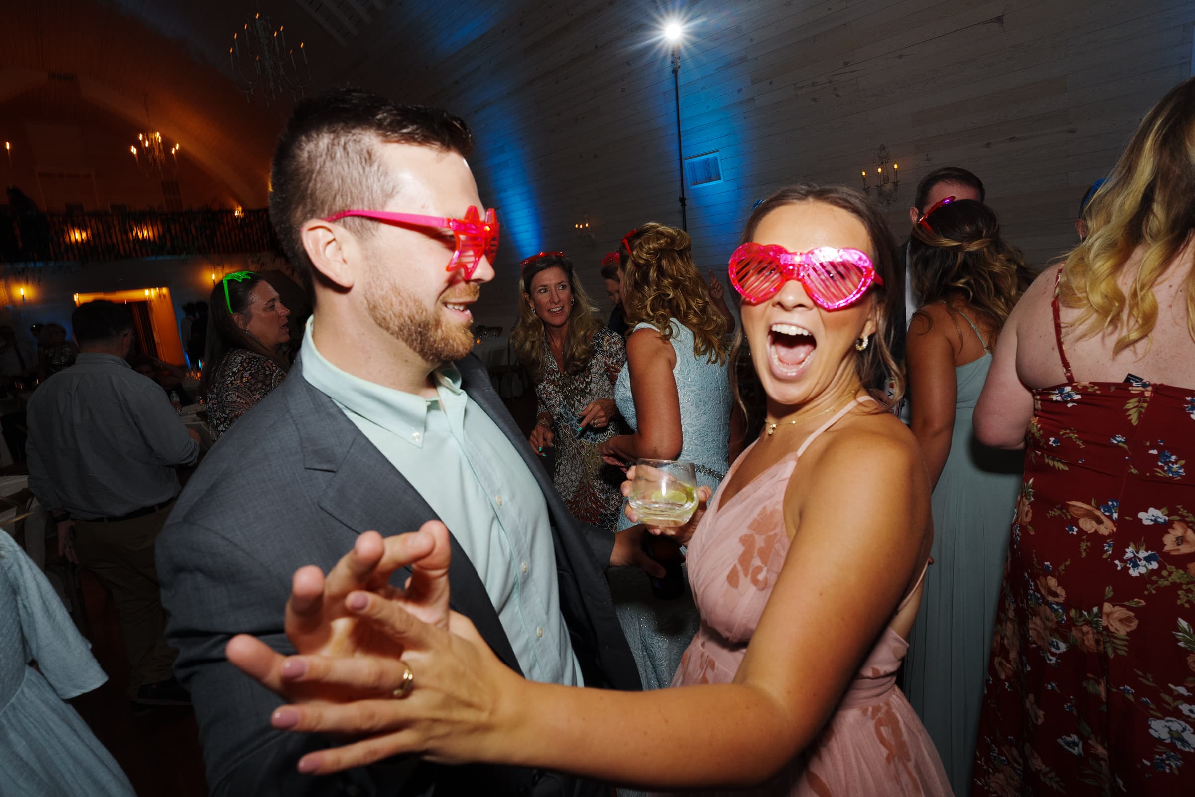historic wakefield barn wedding - logan & rhett At a lively Wakefield Barn wedding, a man and woman wearing playful, pink, heart-shaped sunglasses dance energetically. The woman holds a drink and smiles widely as guests revel in the festive atmosphere under dim lighting.