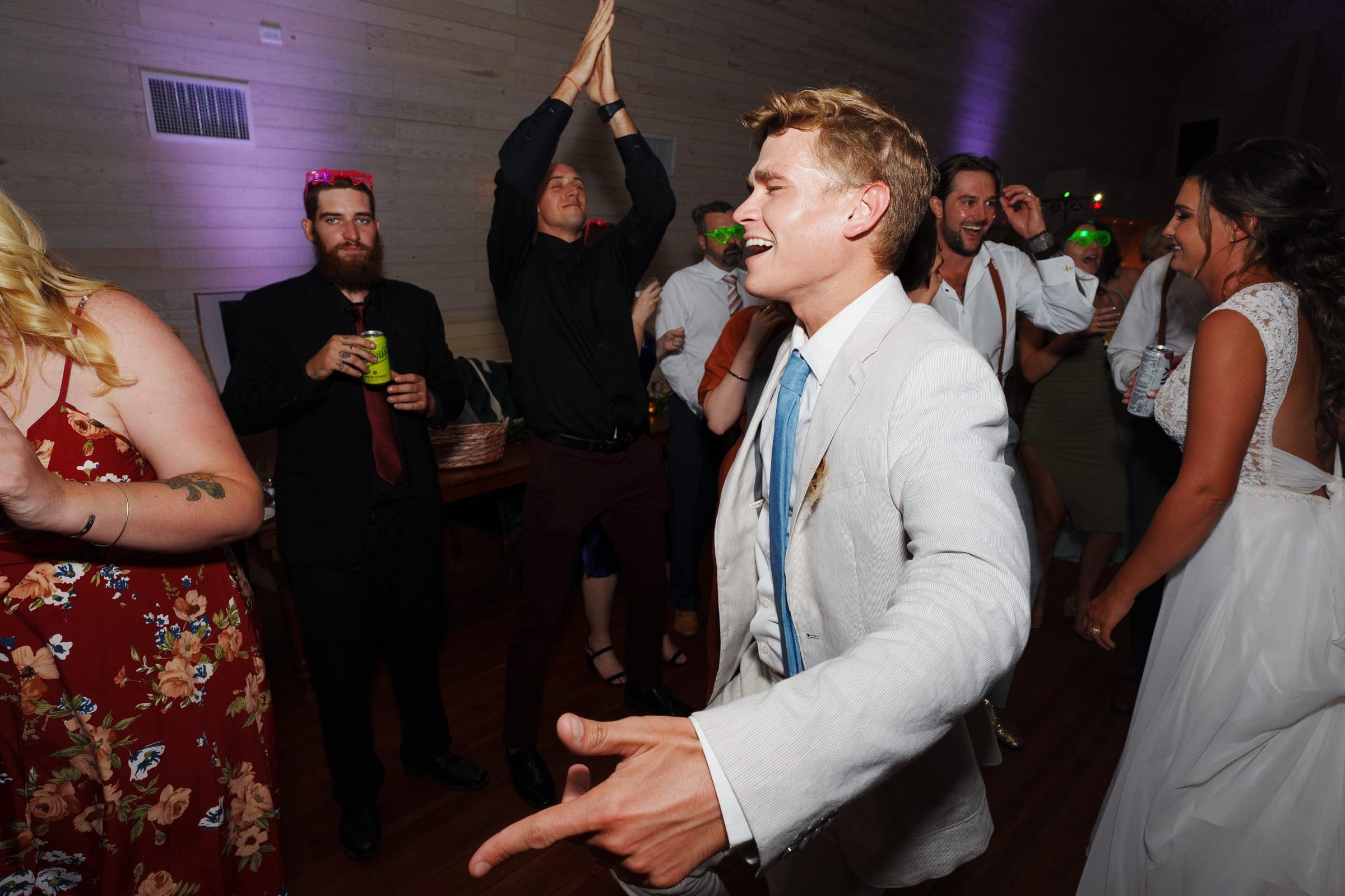 historic wakefield barn wedding - logan & rhett The lively Wakefield Barn wedding reception buzzes with energy as people dance and celebrate. A man in a light-colored suit dances energetically in the foreground, while others clap and enjoy the festivities. Guests, dressed in formal attire, add to the joyous atmosphere.
